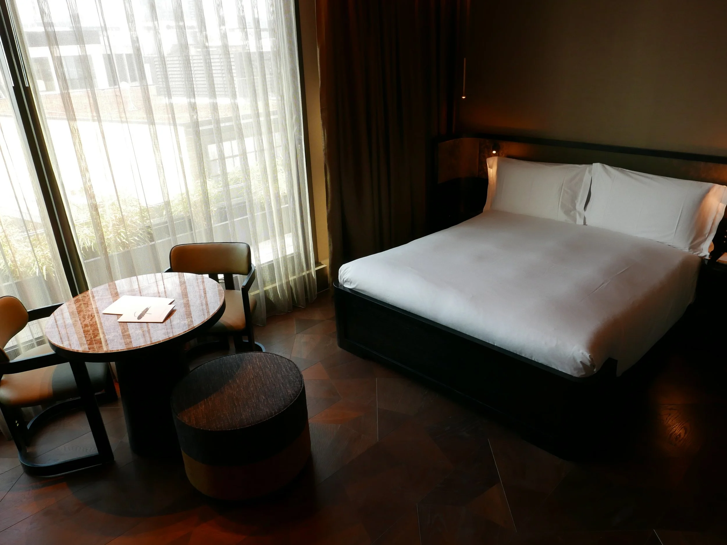 A hotel room with a bed, a round dining table with chairs, and large window with sheer curtains. Nobu Shoreditch roof extension