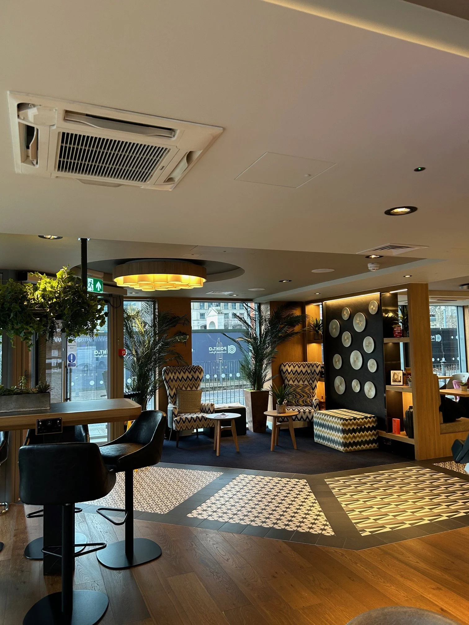 Interior of a modern lounge area with wooden flooring, seating chairs with zigzag patterns, and potted plants near large windows. There is a decorative wall with circular accents and a yellow circular ceiling light. Point A Hotel Canary Wharf