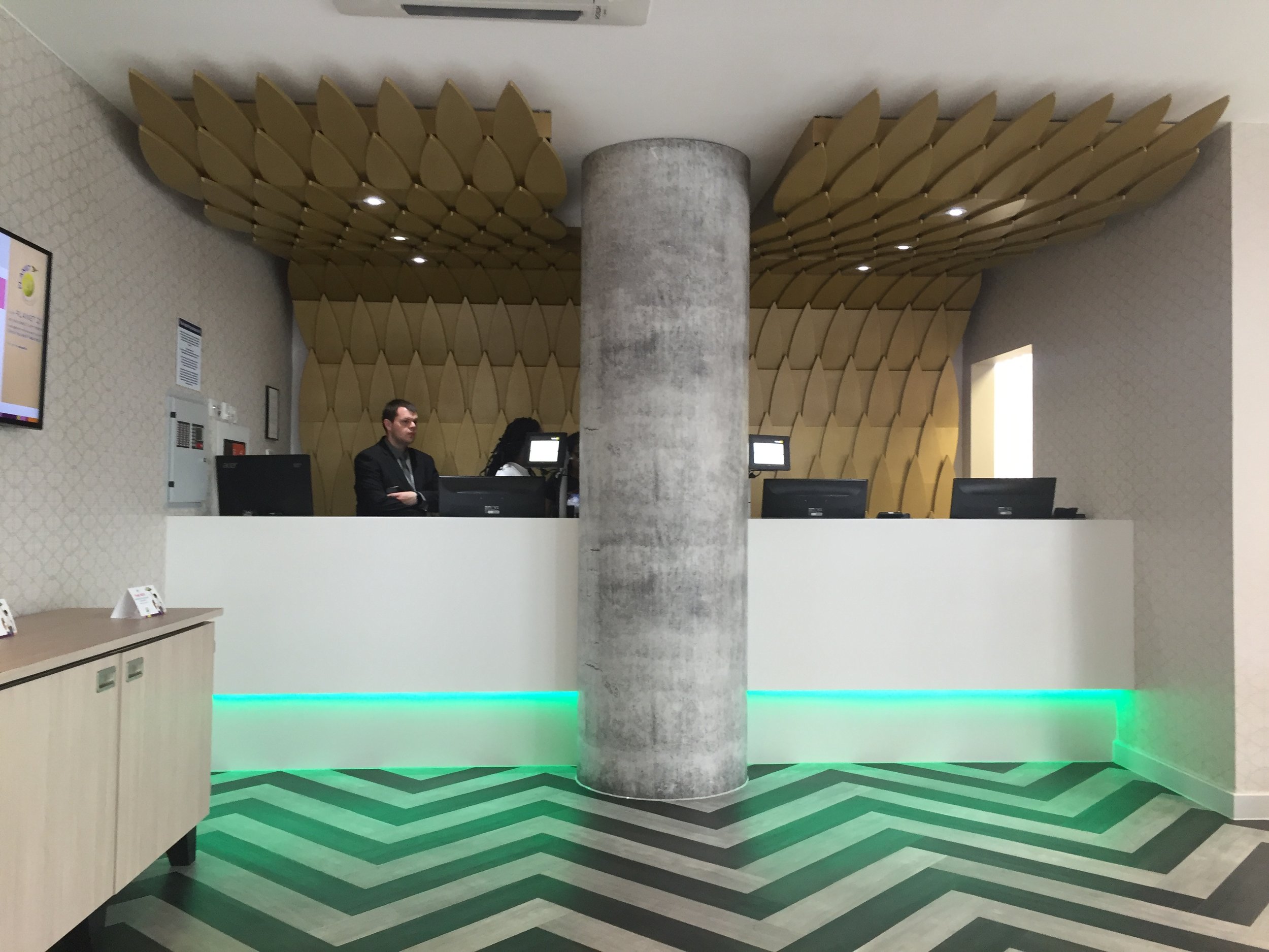 Modern reception area with a large central concrete pillar, a chevron-patterned green and gray floor, and a decorative gold ceiling of the Ibis Style Hotel Bath Road