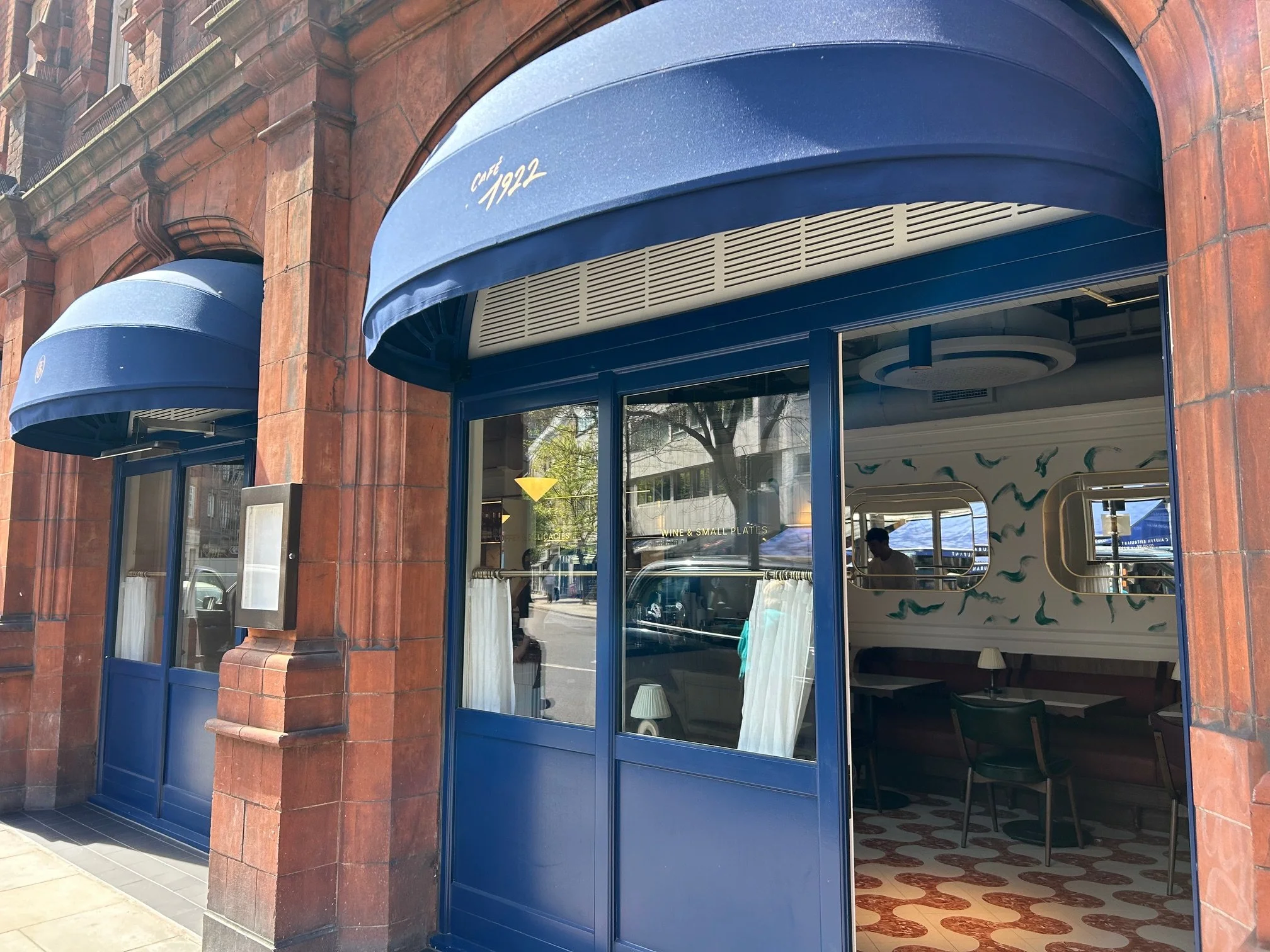 The storefront of a restaurant or cafe with blue awnings, located in a red brick building, with the words 'Cafe 1922' written on the awning, and large glass windows revealing the interior. The Slone Club Café 1992