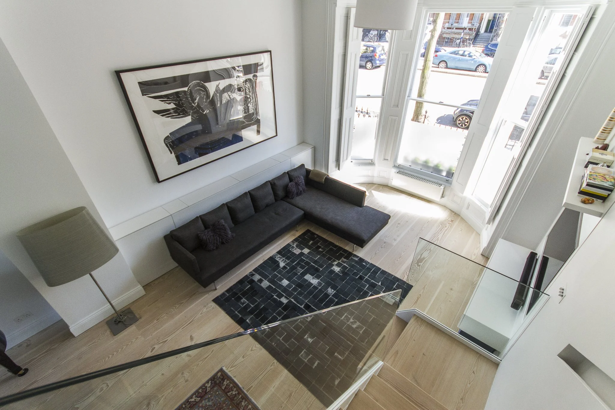 View of the living room from the staircase,  large windows, black sofa with purple pillows, white walls  and Dinesen wooden floor, and a tall floor lamp. Sutherland Avenue House