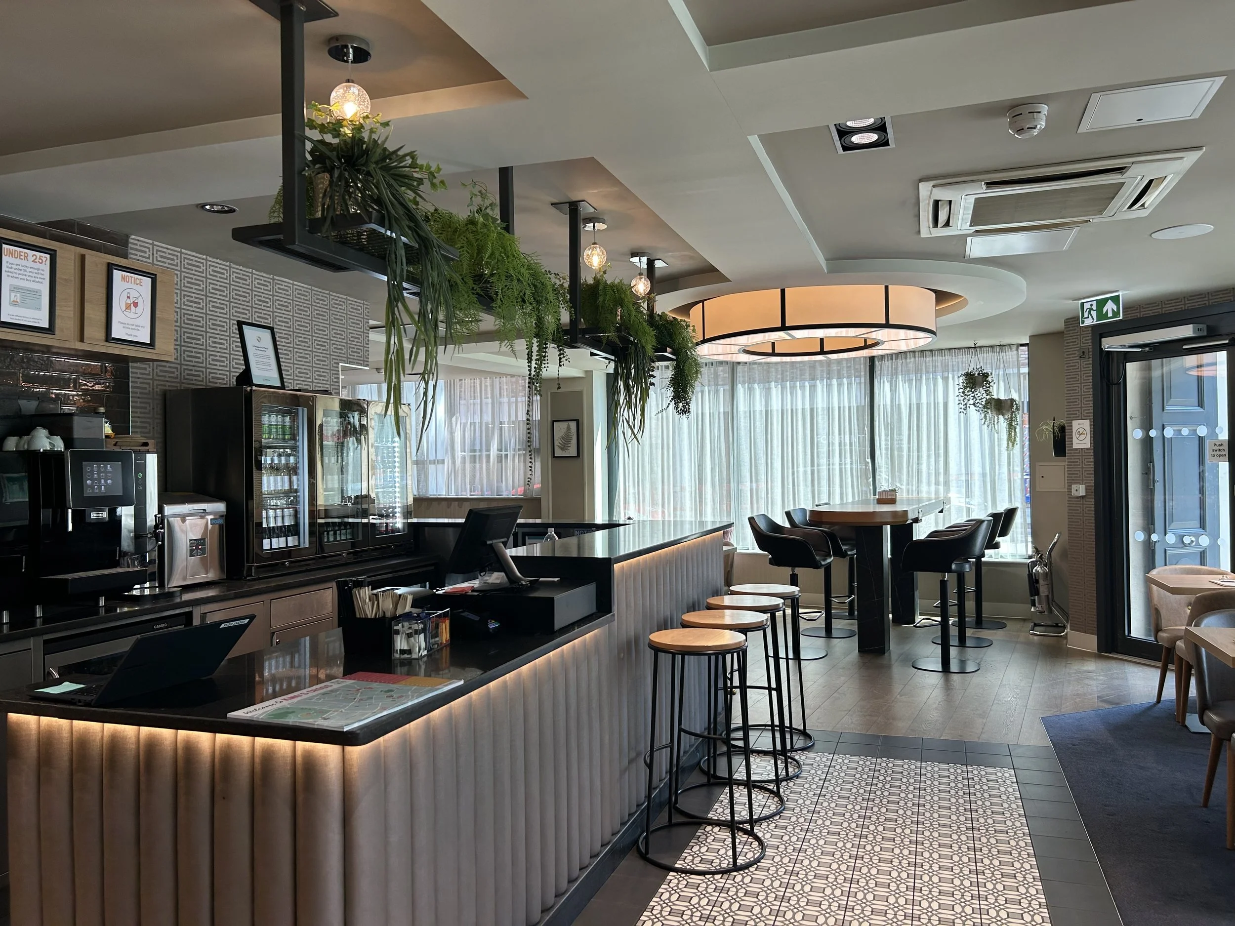 Reception and bar with hanging green plants, a circular light fixture, tables and chairs, and large windows with sheer curtains letting in natural light. of the Point A Hotel Kings Cross