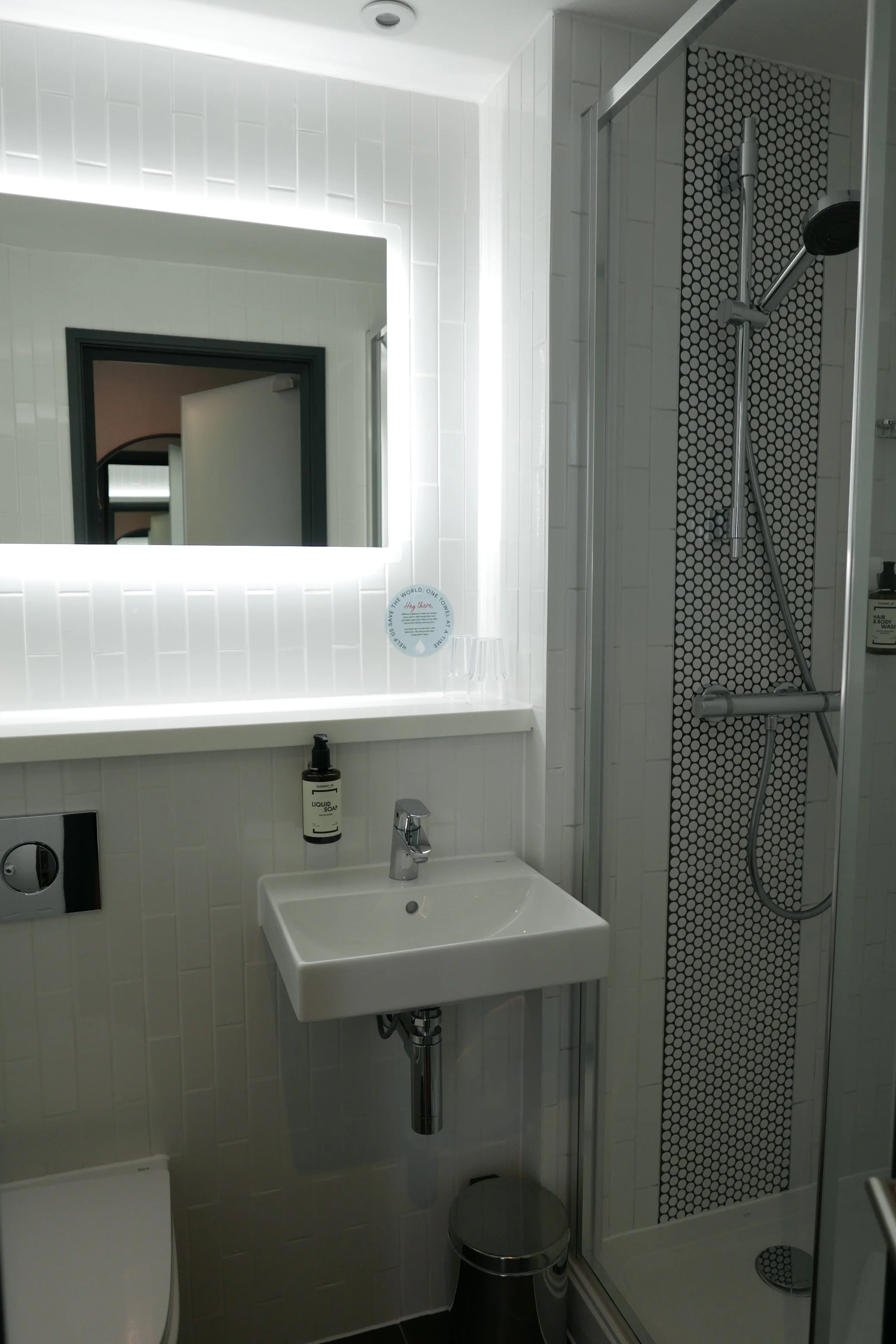 Small modern bathroom with a white sink, a mirror with backlighting, a shower with a glass door, black and white hexagon tile, white tiled walls, a grey waste bin, and various toiletries. Point A Hotel Canary Wharf