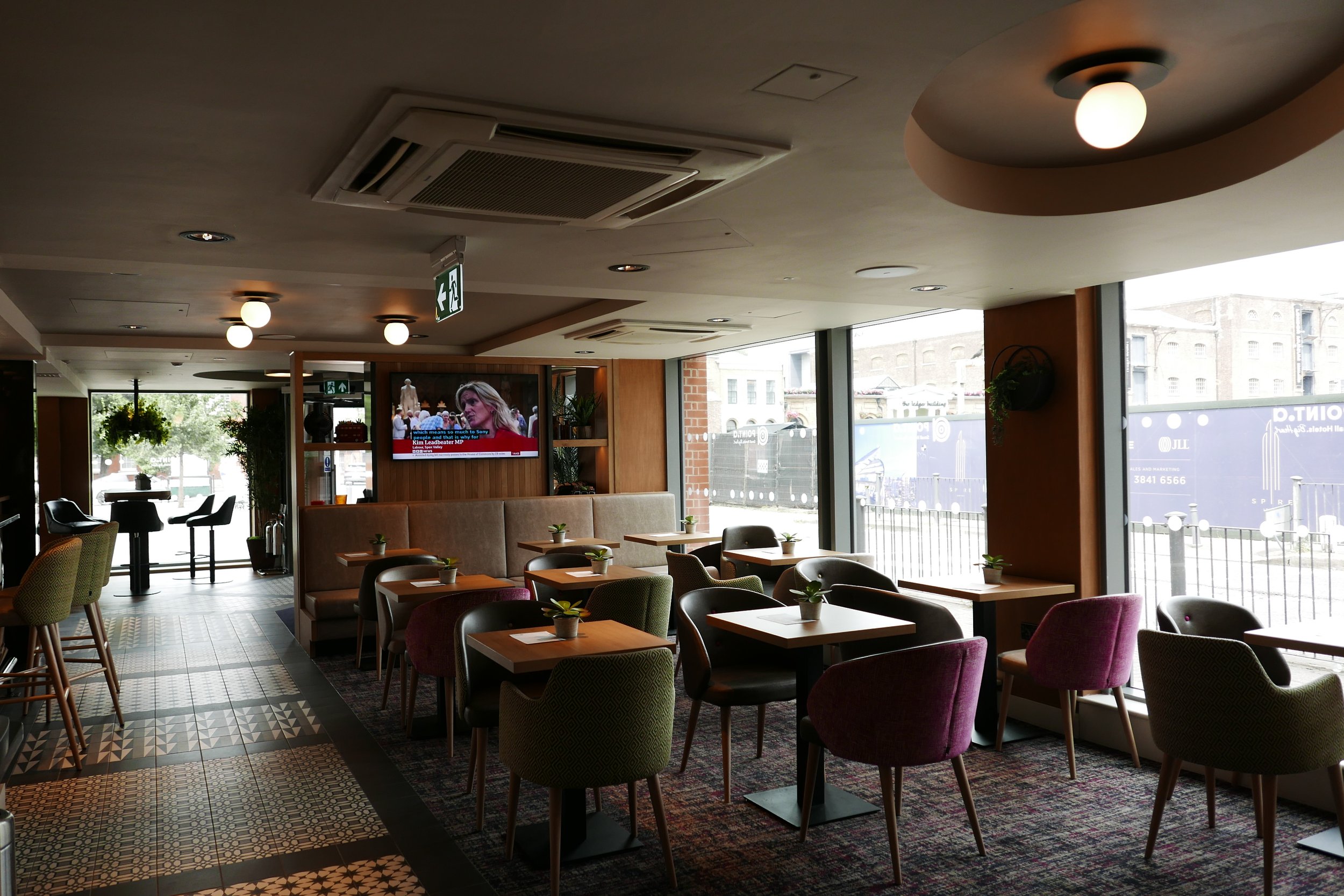Interior of a modern breakfast area with tables and chairs, large windows, TV on the wall, and ceiling lights. Point A Hotel Canary Wharf