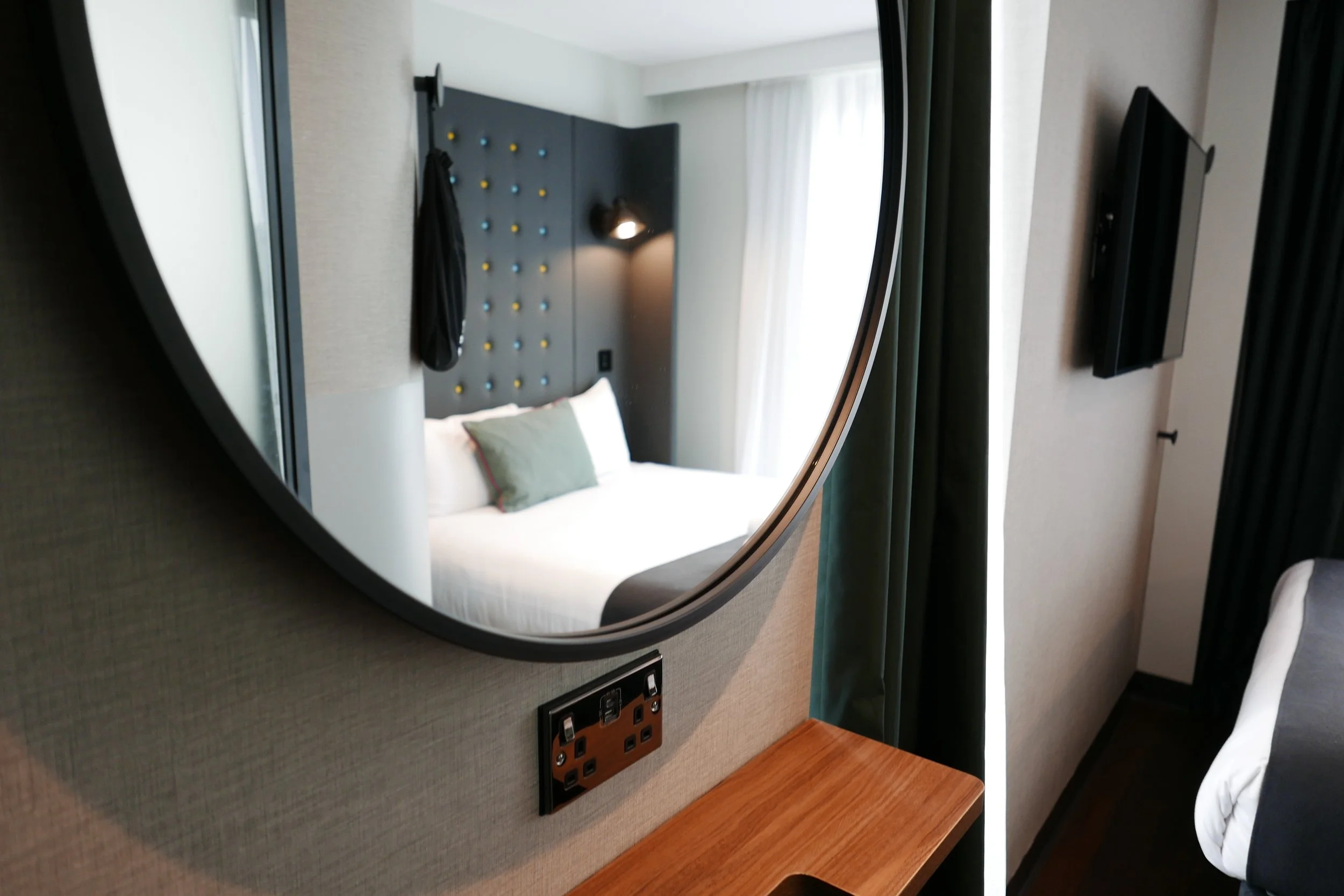 A hotel room mirror reflects a bed with two white pillows, a black headboard with colorful push pins, a wall-mounted lamp, a flat-screen TV, and curtains by a window. Point A Hotel Canary Wharf