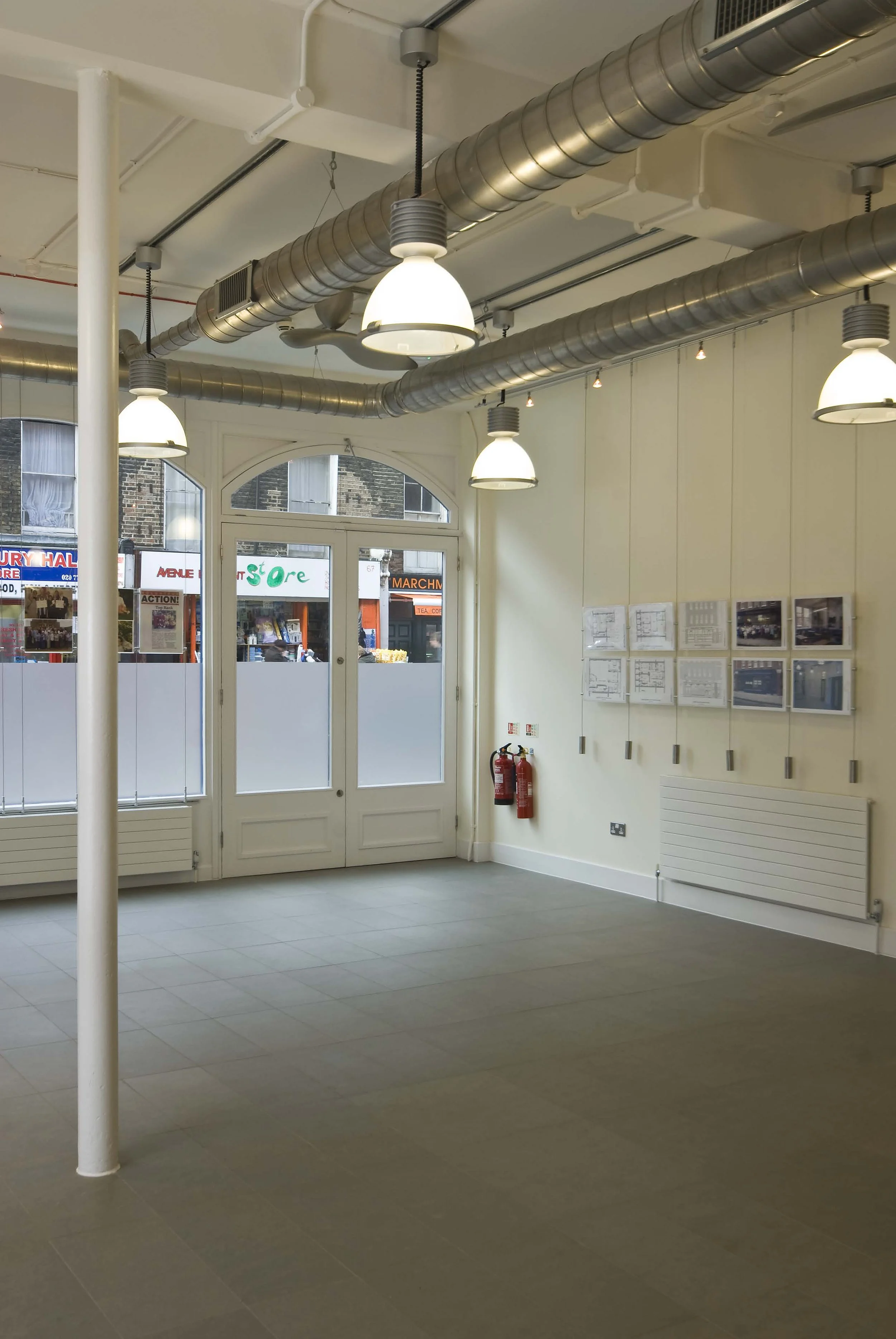 Interior of the main multi purpose room with a white door and large windows, exposed ductwork and hanging lights on the ceiling, photographs or plans on the wall. Marchmont Community Centre