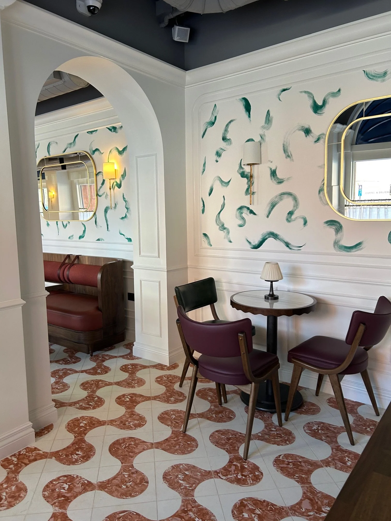 A corner of a restaurant or cafe with a small round table and four maroon chairs. The wall has green squiggle patterns, and there are two wall-mounted lamps with yellow shades. An archway and mirror are visible, along with a patterned tiled floor in 