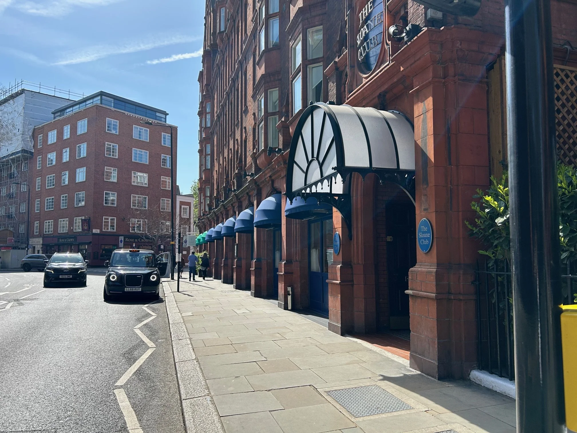 City street scene with a brick building labeled 'The Sloane Club', storefront with blue awnings, parked cars, pedestrians, and a modern high-rise building in the background under a blue sky. The Slone Club Café 1992