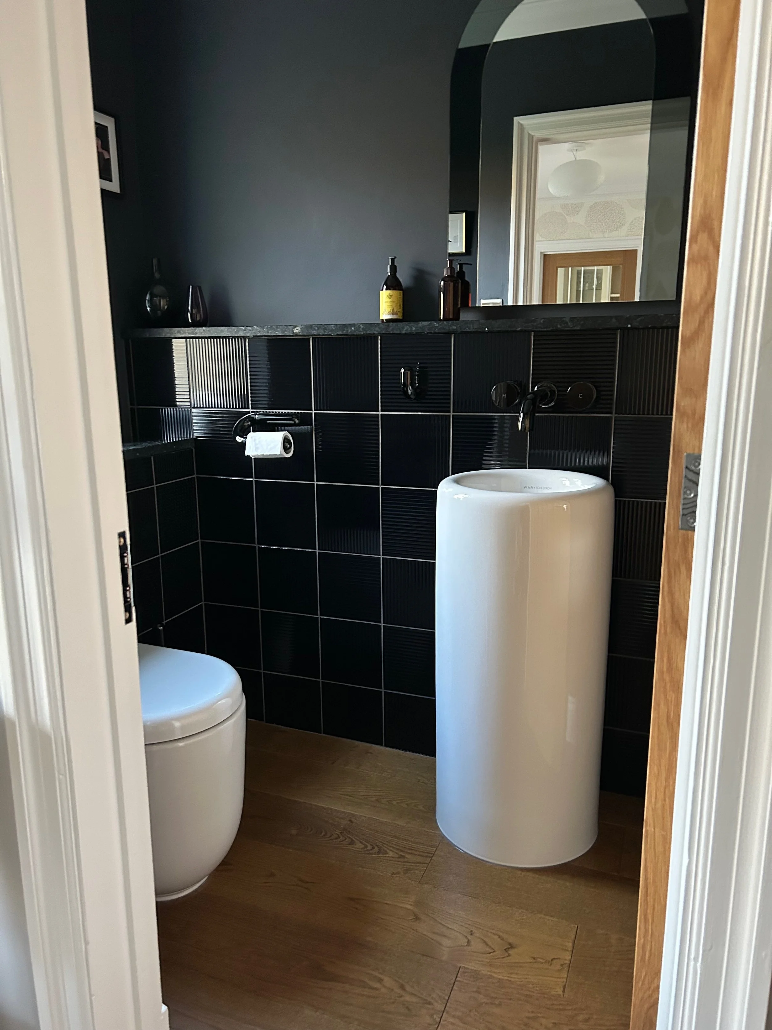 A small bathroom with black tiled walls, a white toilet, and a white cylindrical standalone sink. There is a dark countertop with bottles on it, a paper towel roll, and a large mirror reflecting the ceiling and a doorframe to another room. The bathro