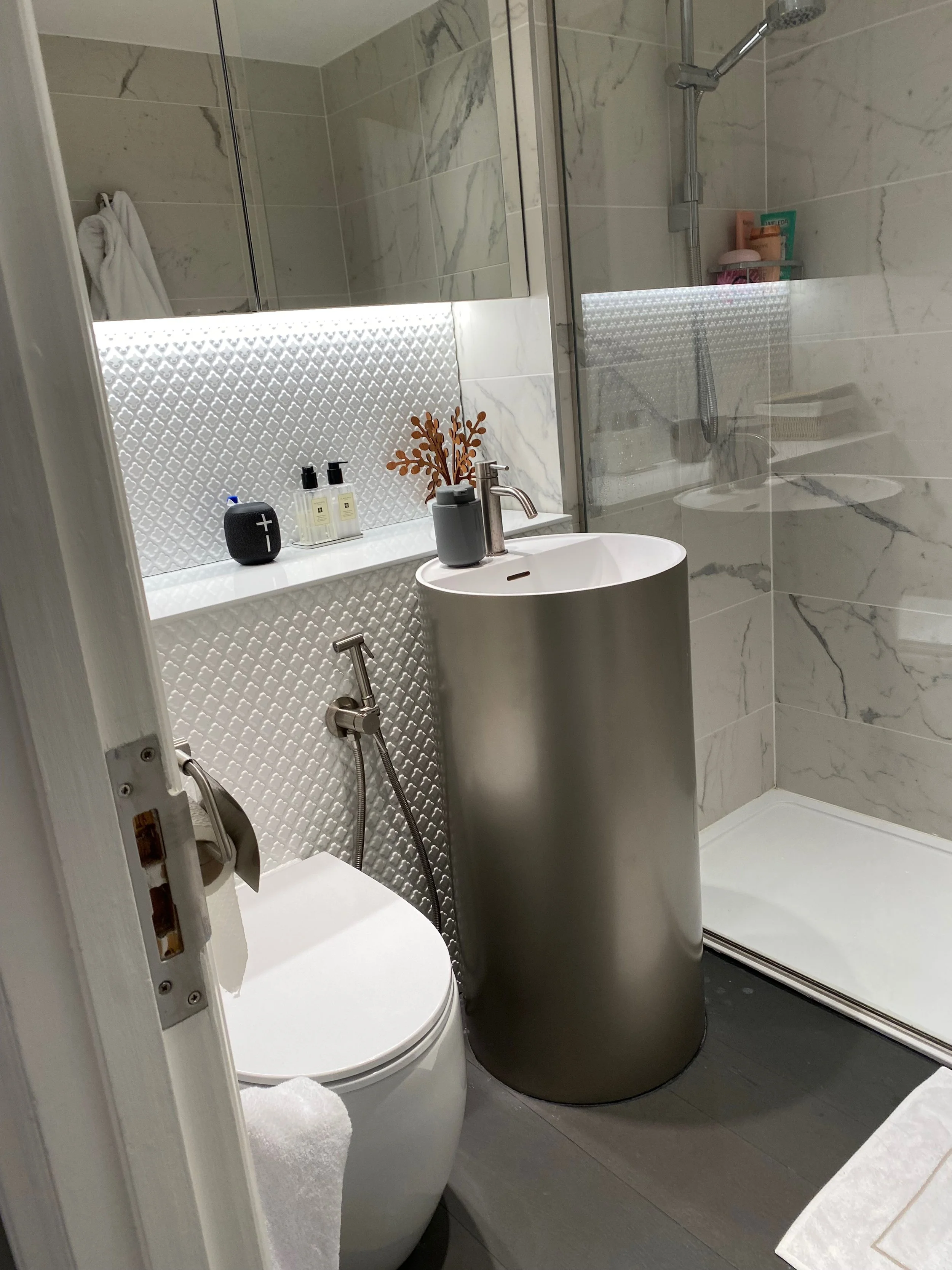 A modern bathroom with a walk-in shower, a white bidet, and a small white sink with a metal faucet. There are toiletries on a shelf inside the shower and a decorative vase with dried branches on the sink. The walls are covered with marble tiles, and 