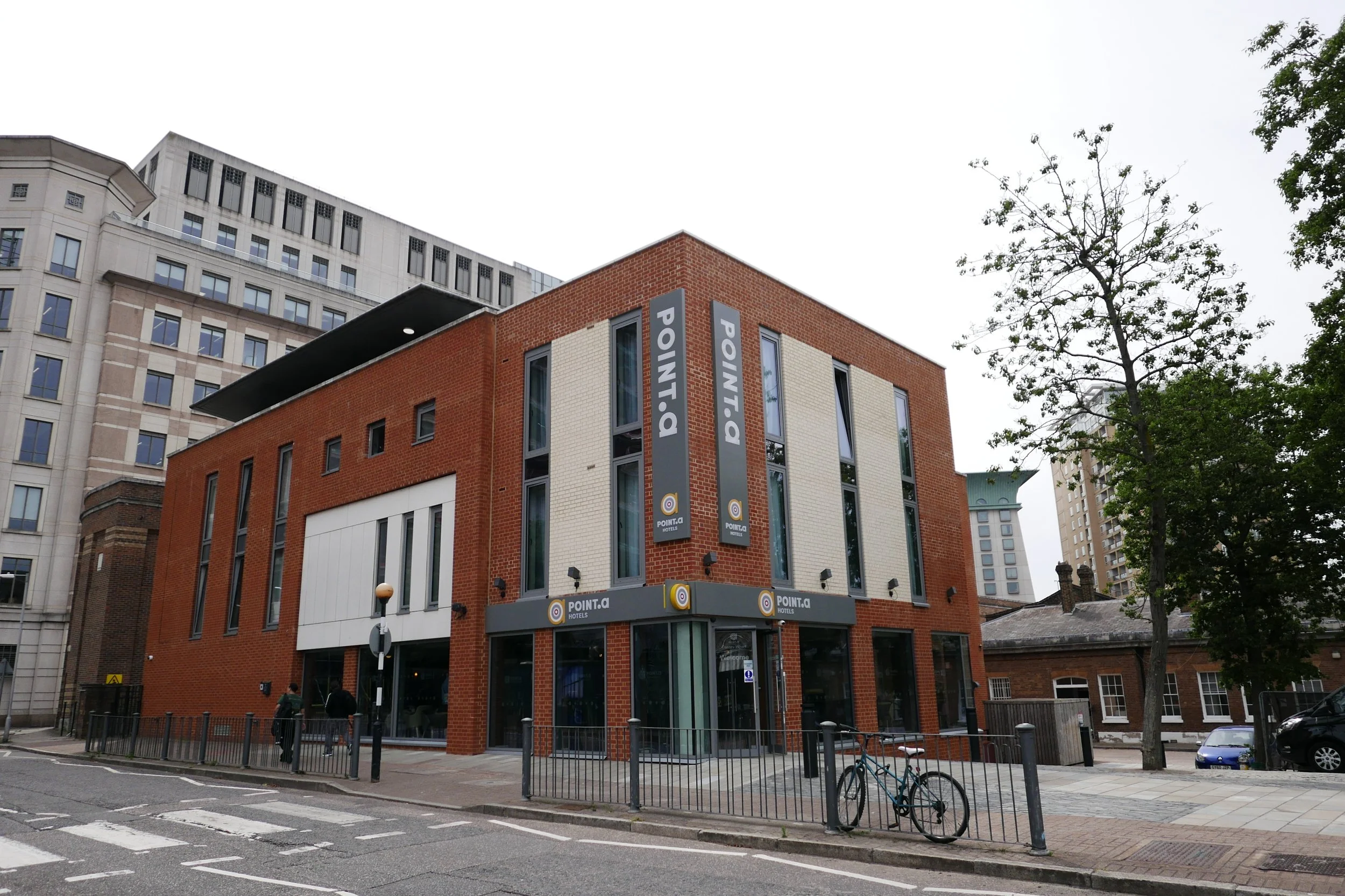 Street view of a modern multi-story brick building with 'POINT A' signage, adjacent to high-rise buildings, trees, and a bicycle parked on the sidewalk. Point A Hotel Canary Wharf