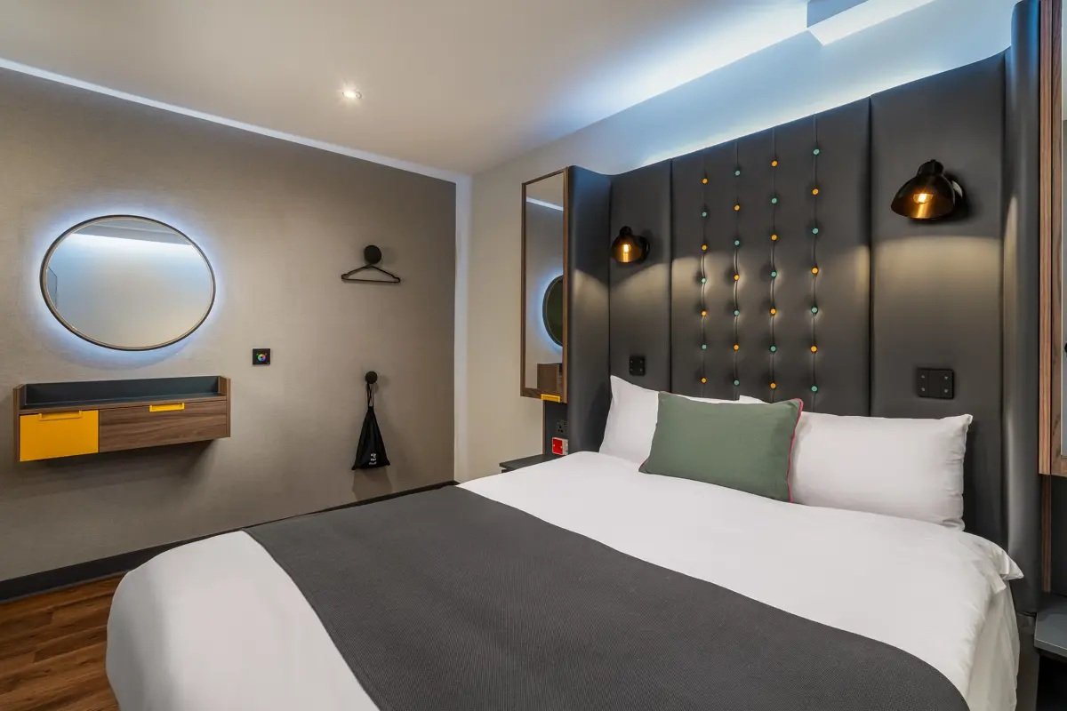 Modern hotel bedroom with a large bed, decorative headboard with colored lights, wall-mounted lamps, a mirror of the Point A Hotel Shoreditch