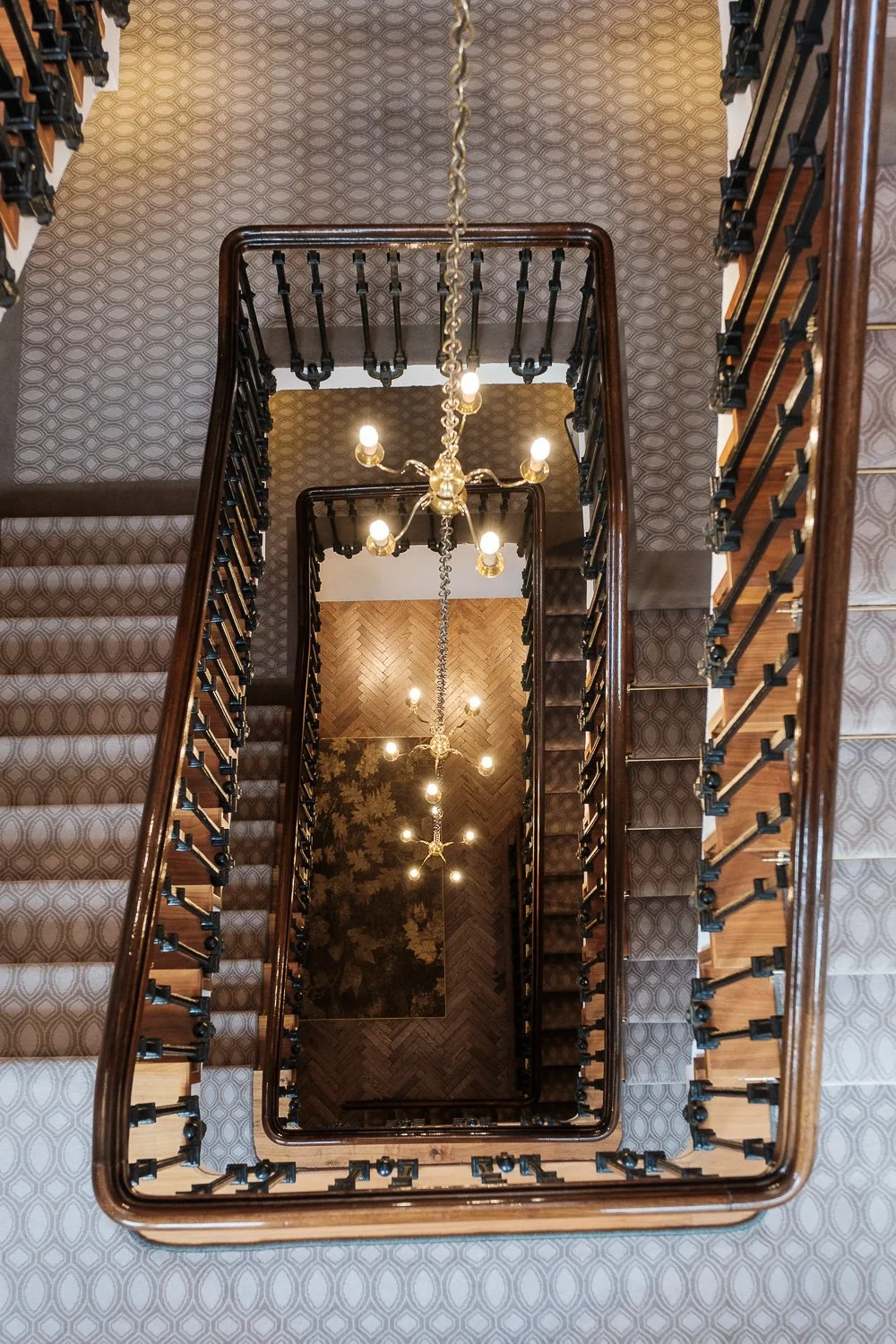 Looking down at a multi-story wooden staircase with patterned carpet, with chandeliers hanging from the ceiling. Sloane Place Restaurant