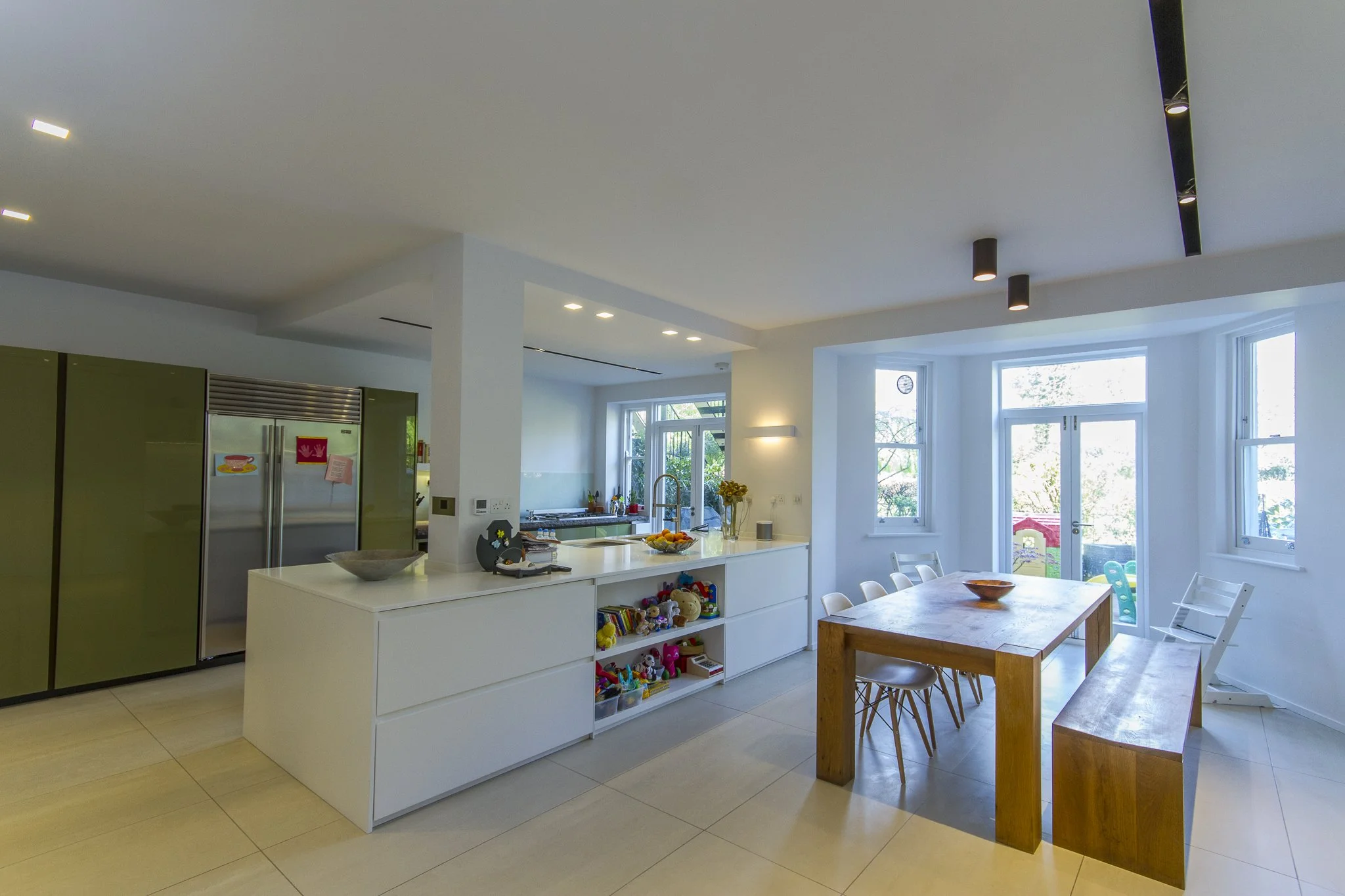 A modern open-concept kitchen and dining area with large windows, a white kitchen island, a wooden dining table with white chairs, and a ceiling with recessed and track lighting. There is a refrigerator and various kitchen items visible. Sutherland A