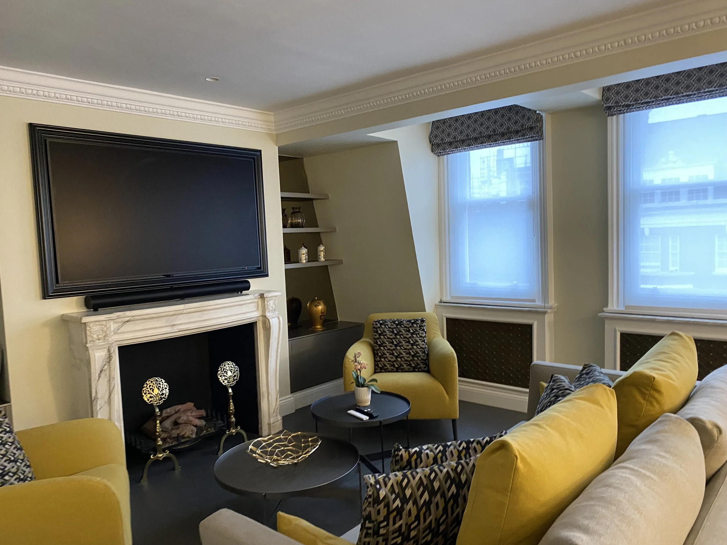 Living room with yellow armchairs, black round coffee tables, a fireplace with a white marble mantel, and a flat-screen TV mounted above it. Large windows with patterned valances let in natural light. Pont Street Knightsbridge 