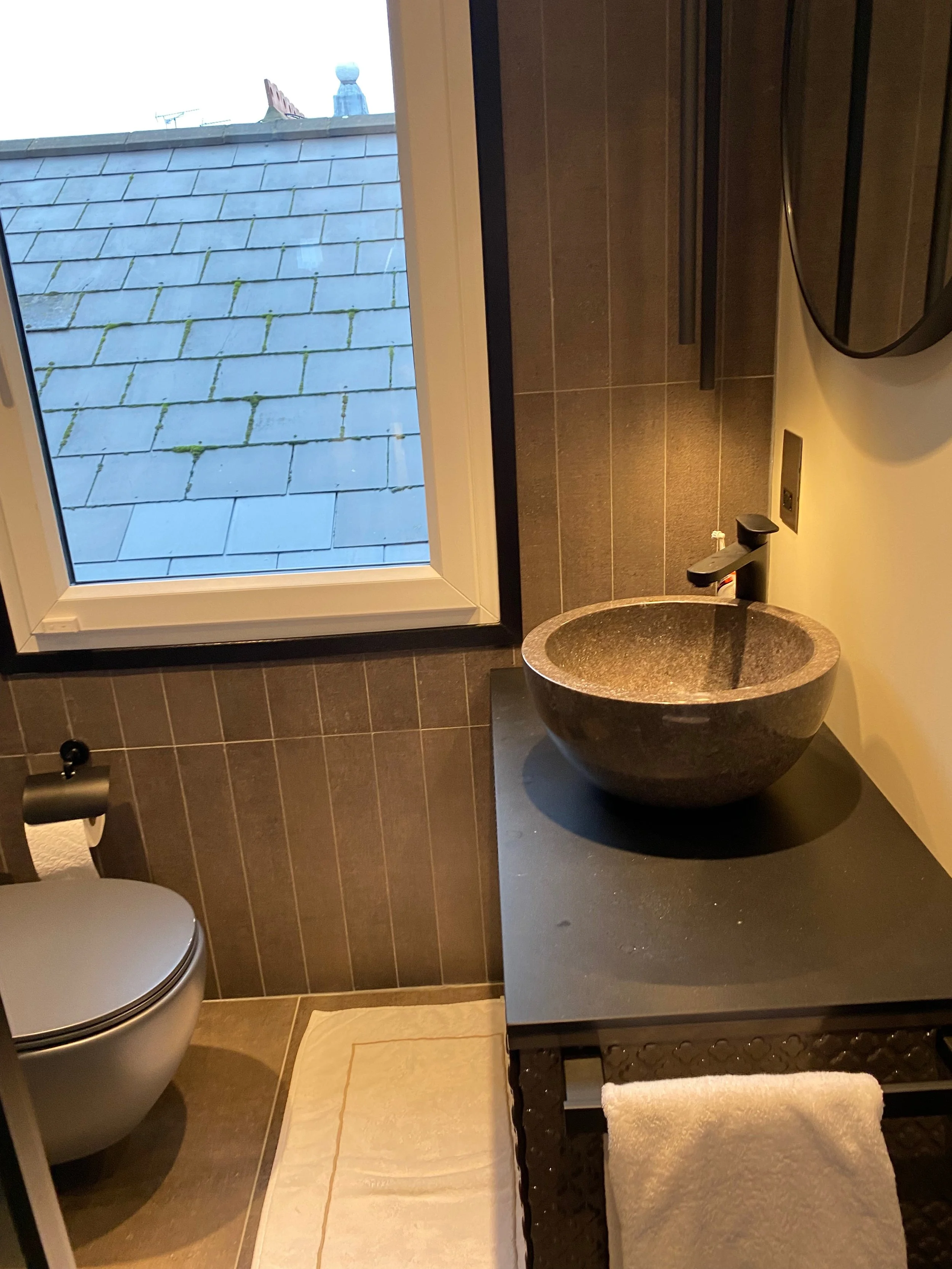 A small bathroom with a window showing a tiled roof outside. The bathroom has a white toilet, a black sink with a granite bowl, and a black table with a shelf. There is a towel on the shelf and a toothbrush next to the sink. The floor is tiled, and a