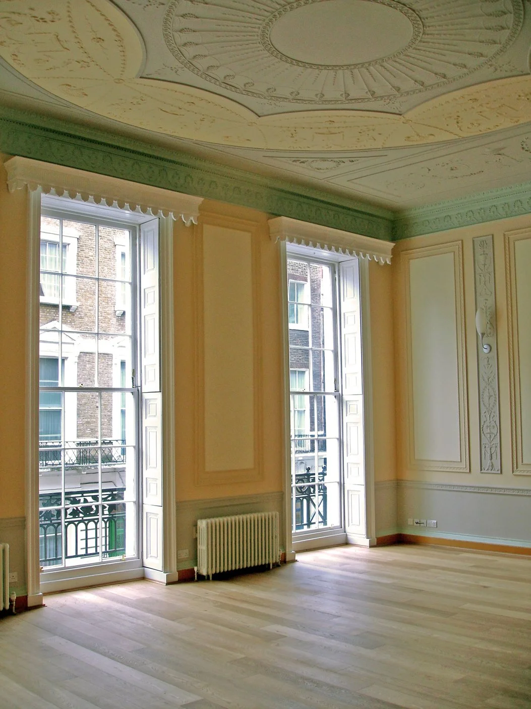 The drawing room room with large windows, ornate ceiling, decorative wall moldings, and hardwood flooring. of the Harley Street apartment in Grade II Listed Building