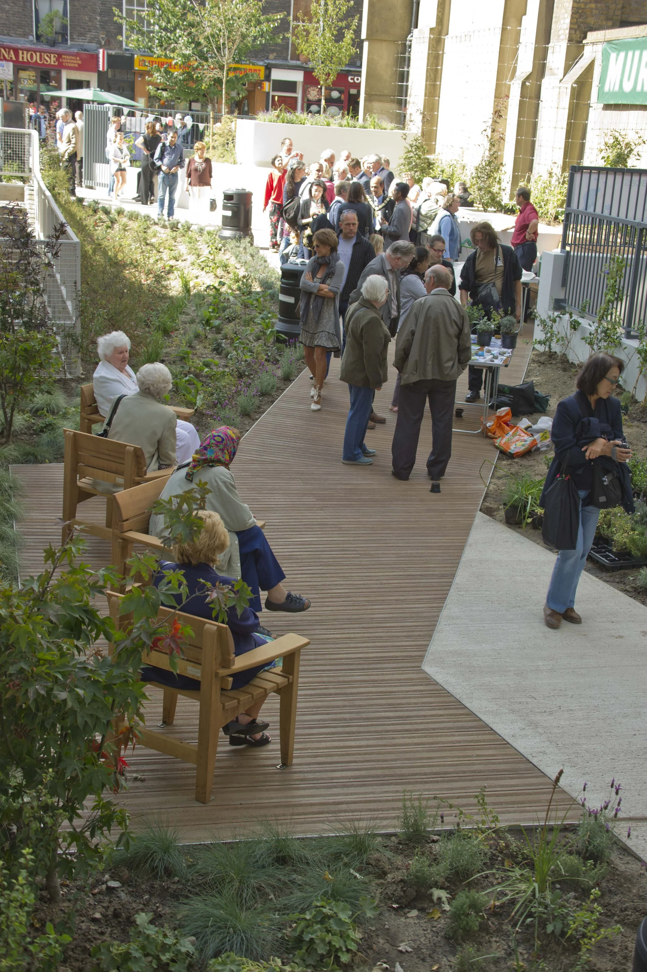View of the central section of the garden with the Millboard decking wooden seating and planting area. A diverse group of people standing and sitting in the garden during the opening day. Marchmont Street Community Gardens