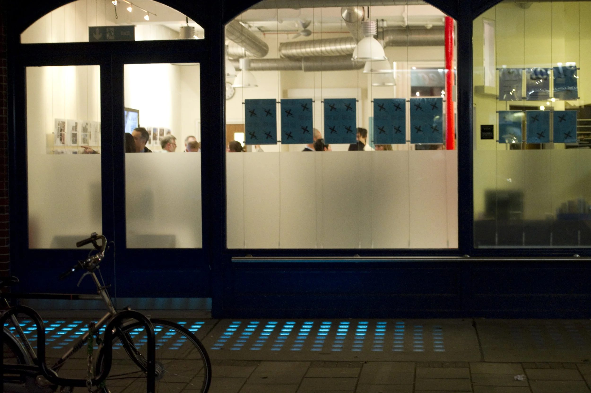 Night view of the exterior of a building with a large window showing people inside and blue posters on the window. There are  illuminated blue ground lights. Marchmont Community Centre