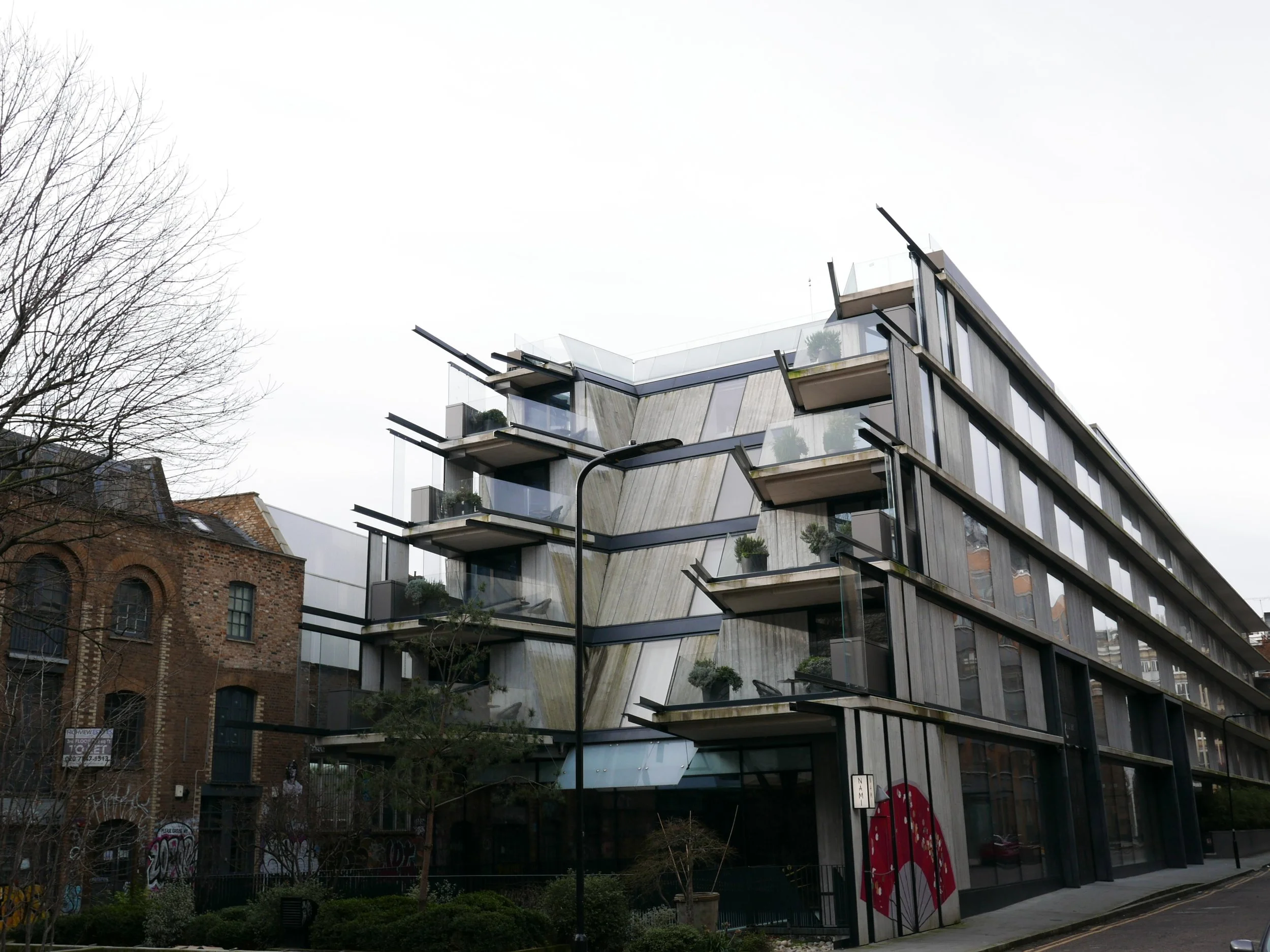 View of the great multi-story Nobu Hotel Shoreditch with glass balconies and greenery, adjacent to a brick building, with bare trees and graffiti in the foreground. Nobu Shoreditch roof extension