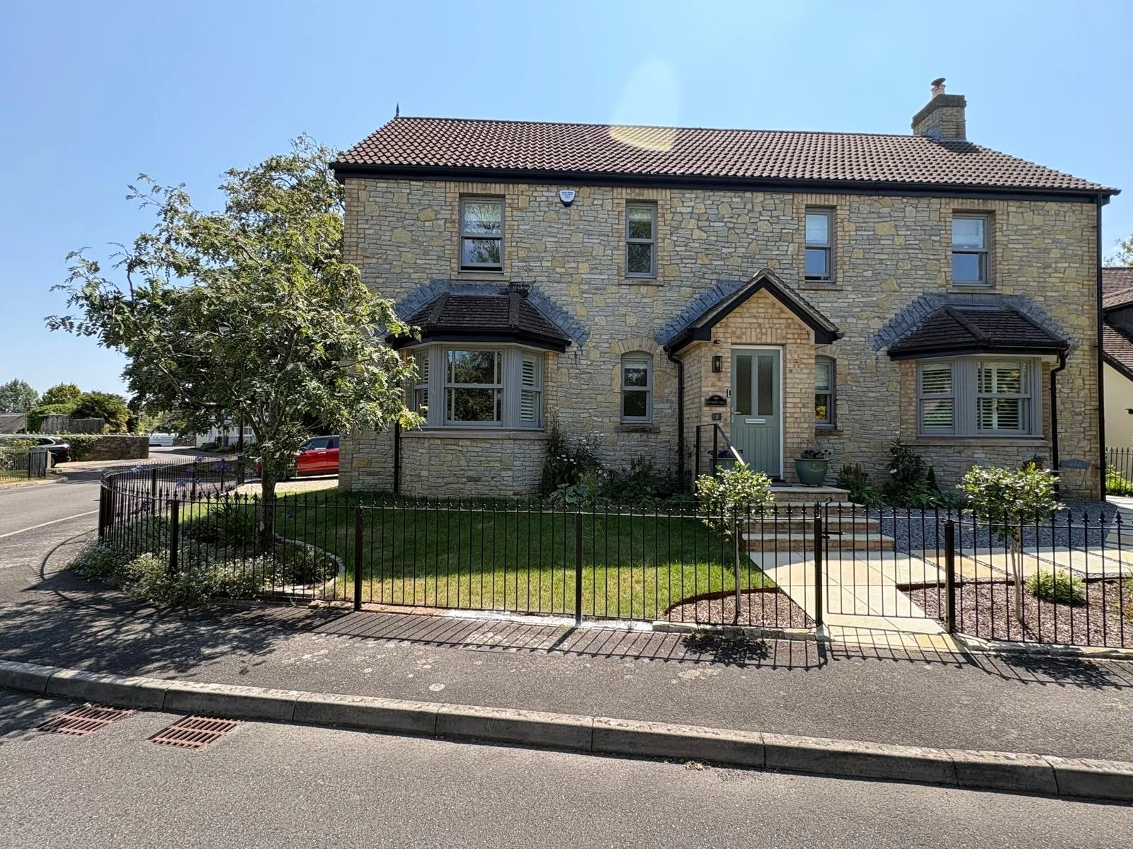 Front view of a two-story house with a stone exterior, front door with steps, bay windows, a small front yard with a bushy tree, and a black metal fence along the sidewalk. Wedmore House North Sommerset 