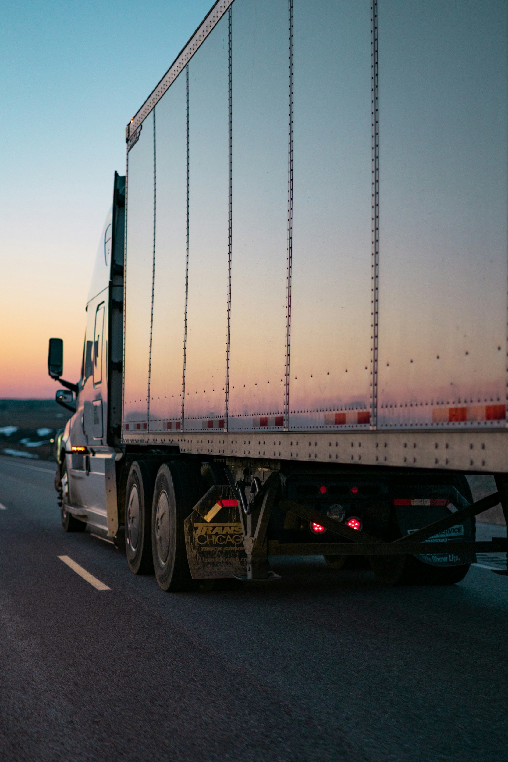 Un camion blanc roulant sur une route au coucher du soleil, le ciel est dégradé entre des teintes rose et bleu.