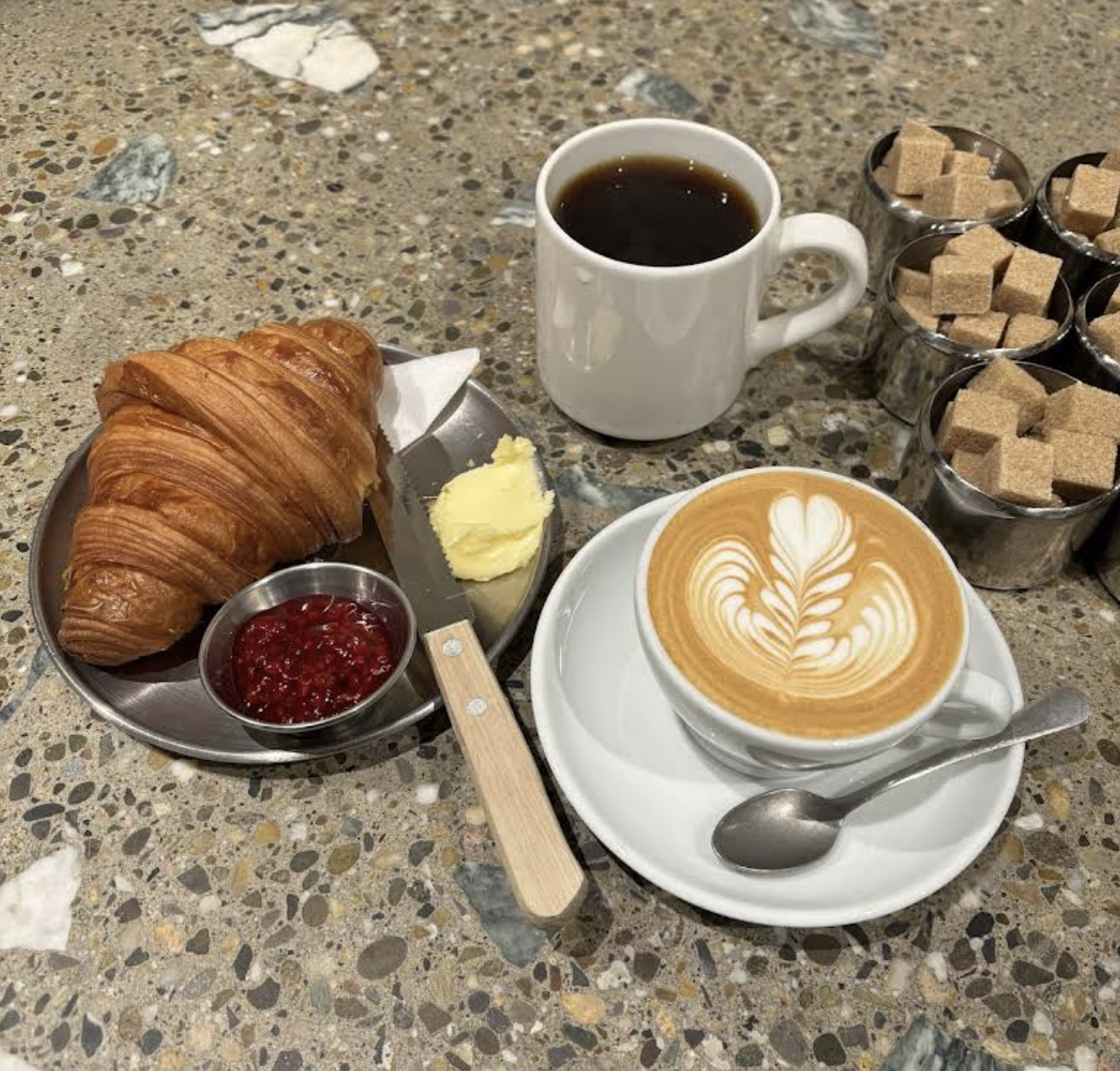A breakfast scene with a croissant, butter, and raspberry jam on a plate, a cup of coffee with latte art on a saucer, a mug of black coffee, and several containers of brown sugar cubes on a speckled granite table.
