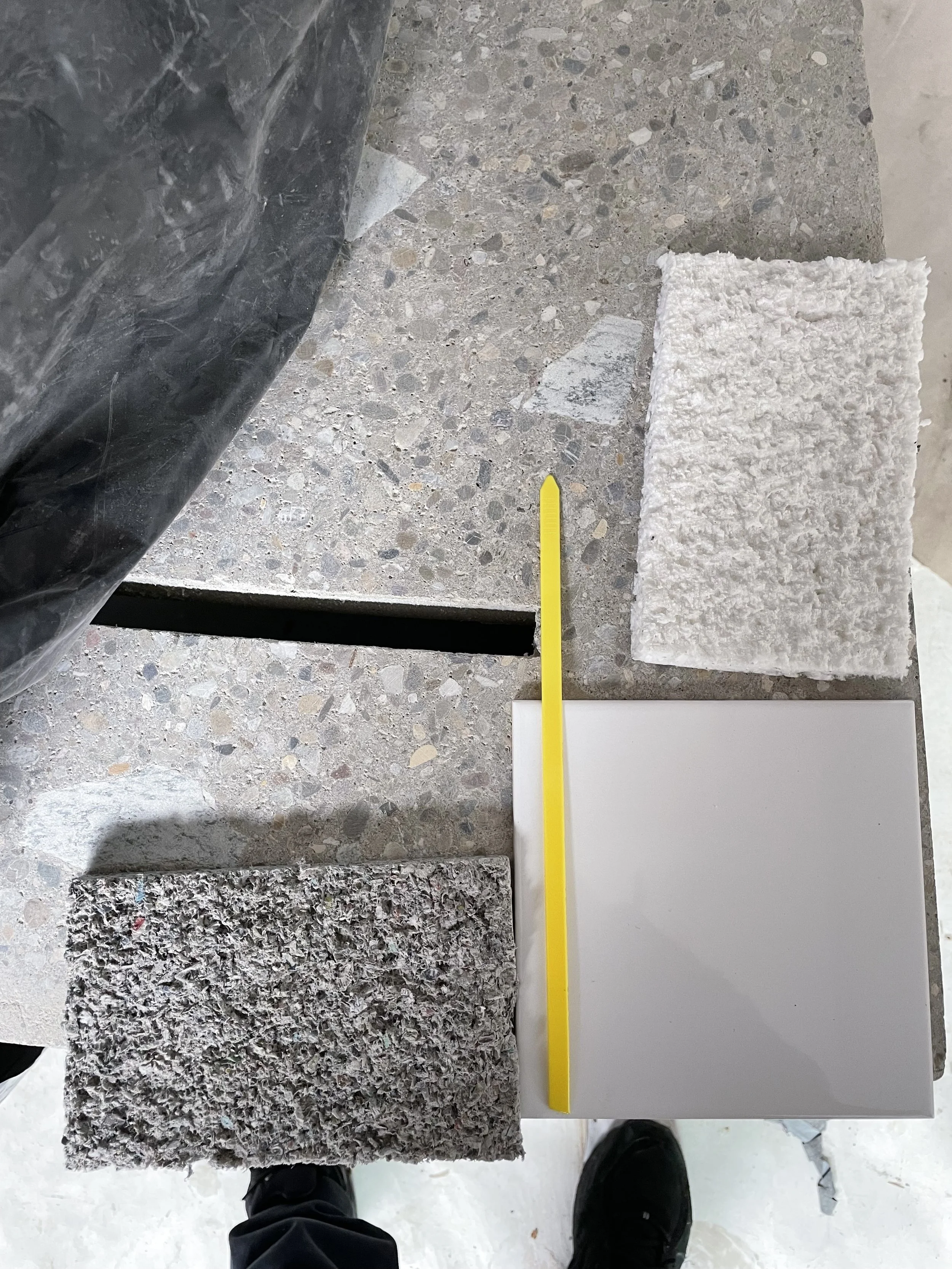 Various sample tiles including granite, marble, and quartz slabs laid on a surface, with a yellow plastic tool and black shoes visible at the bottom.