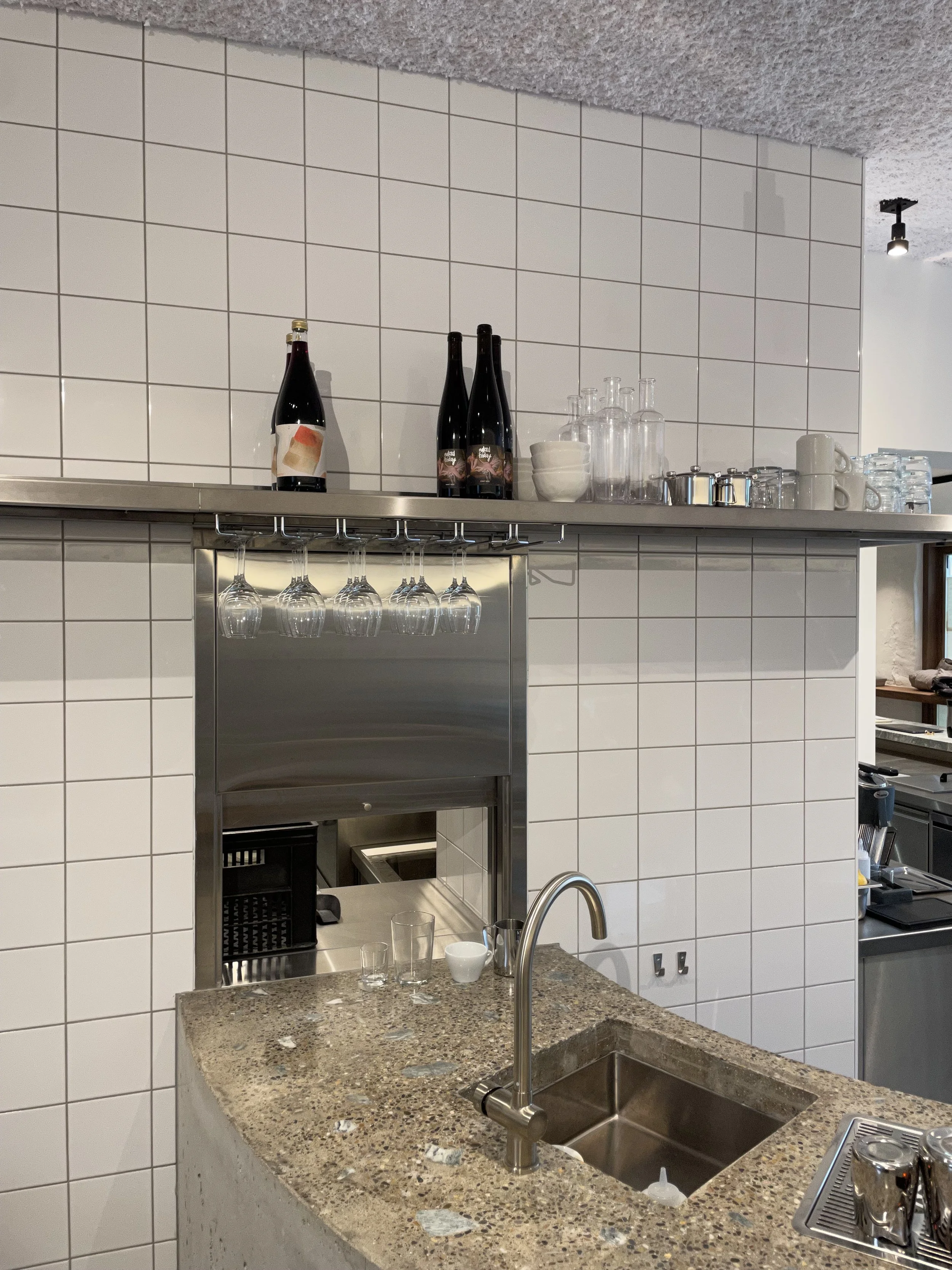 A kitchen with a tiled wall, granite countertop, stainless steel sink, and various bottles, glasses, and mugs on a shelf and countertop.