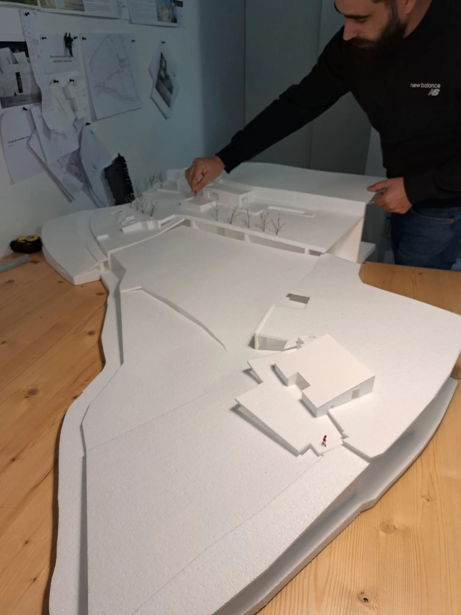A man is working on a scale model of a landscape or architectural project, which includes white foam or styrofoam pieces representing terrain, buildings, and trees. He is leaning over the model, placing or adjusting an element, with a wall behind him