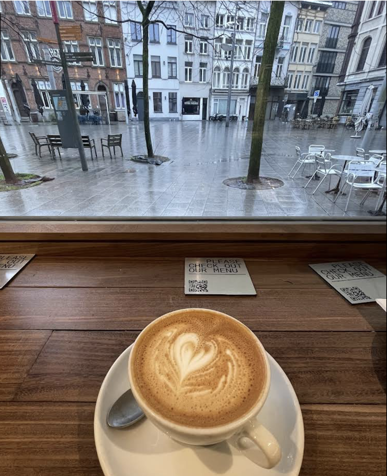 Indoor cafe view through a window with rain outside, with a coffee cup containing a latte with heart-shaped foam art on a saucer and a spoon, while three small printed menus with QR codes are placed on the wooden table surface.