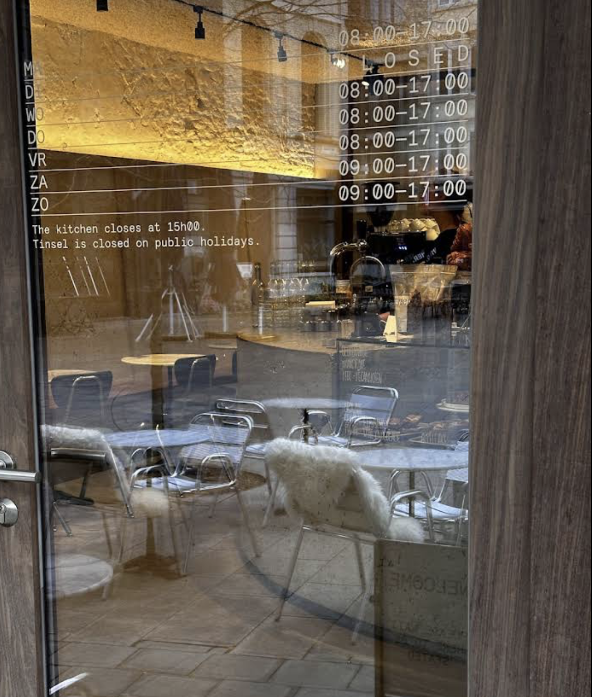 A glass door of a cafe showing the interior with chairs, tables, and a counter. The door has hours of operation and notes about closing times and holidays written on it.