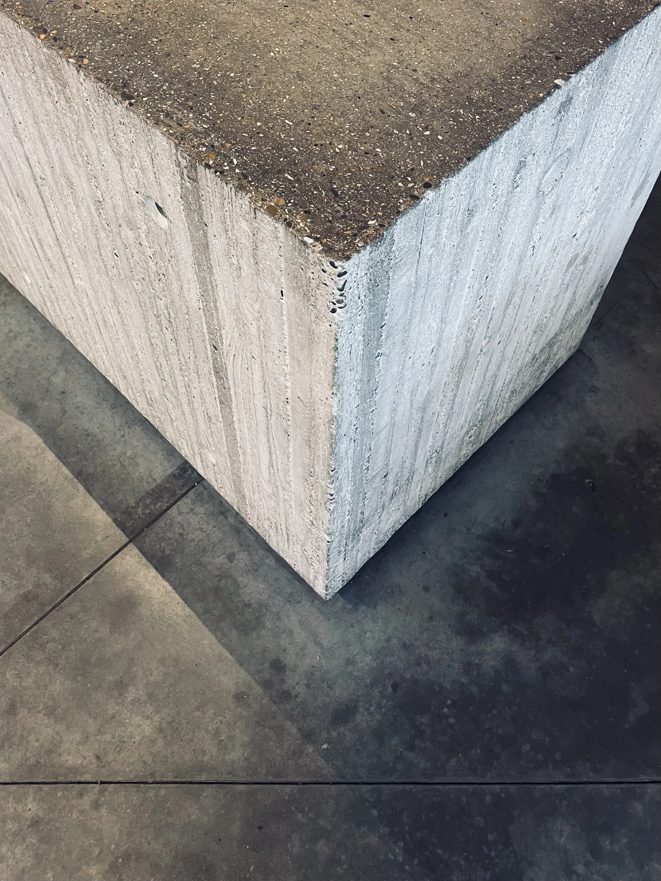 Close-up of a corner of a concrete block with a rough, textured surface. The surface shows small stones embedded in the concrete. The block is placed on a dark, smooth floor.
