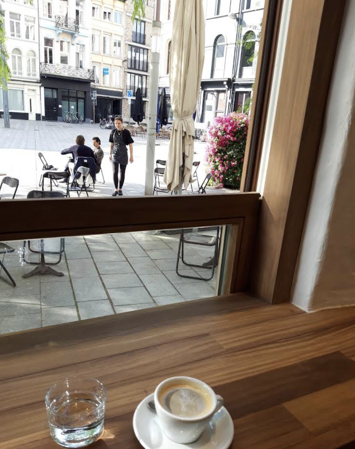 View of an outdoor city plaza seen from a cafe window, with a coffee cup and glass of water on a wooden counter in the foreground.