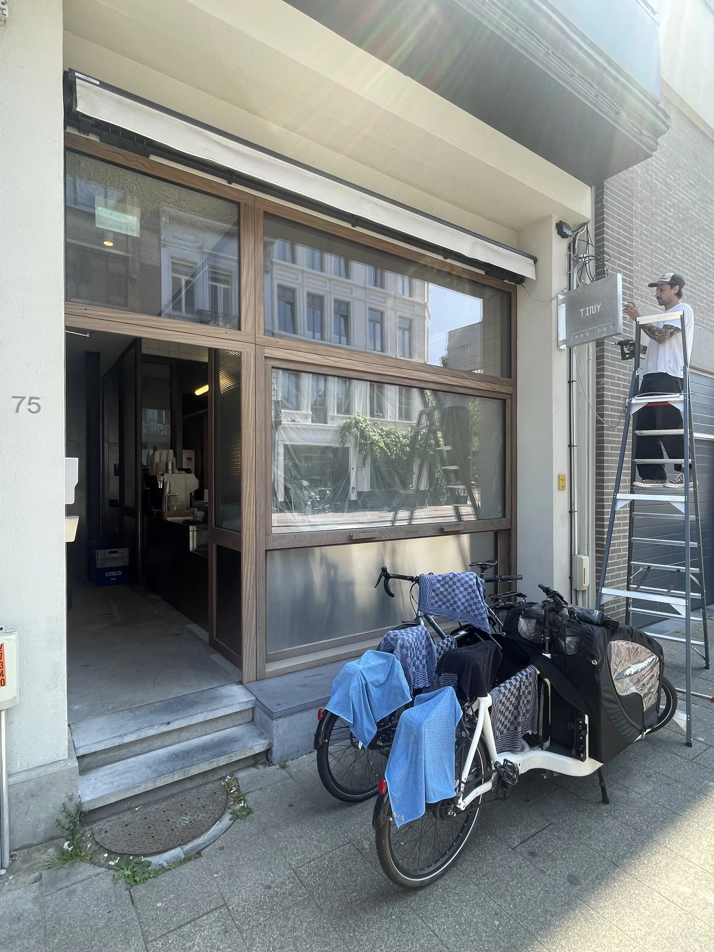A storefront with large glass windows and a wooden frame, two bicycles equipped with delivery boxes and blue rain covers parked outside, and a person on a ladder working near electrical cables.