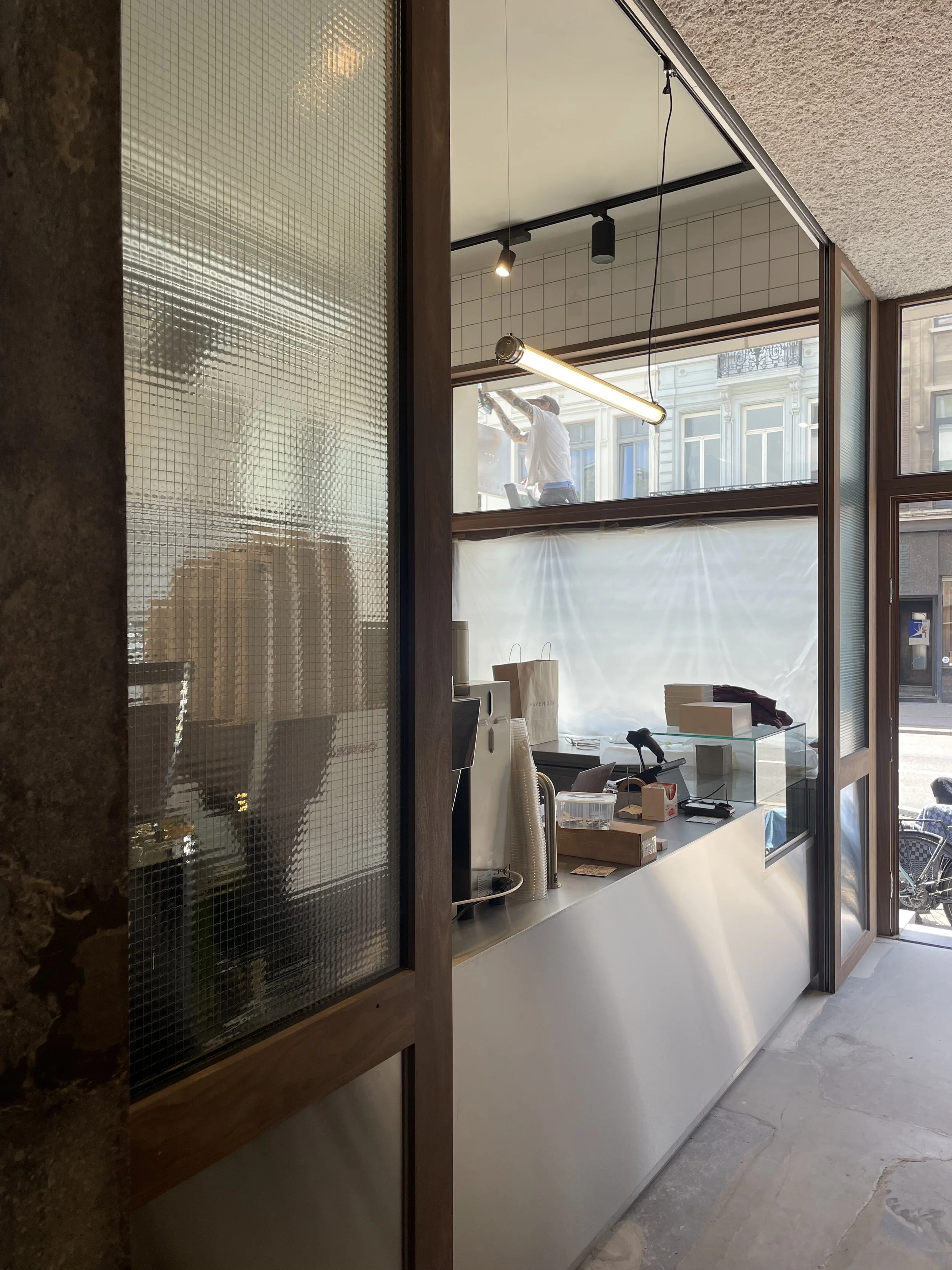 Interior view of a cafe or shop, with a large window and counter area. A person is working outside the window, visible through the glass. Shelves and supplies are on the counter, and a hanging light fixture is above. Textured ceiling and wood accents