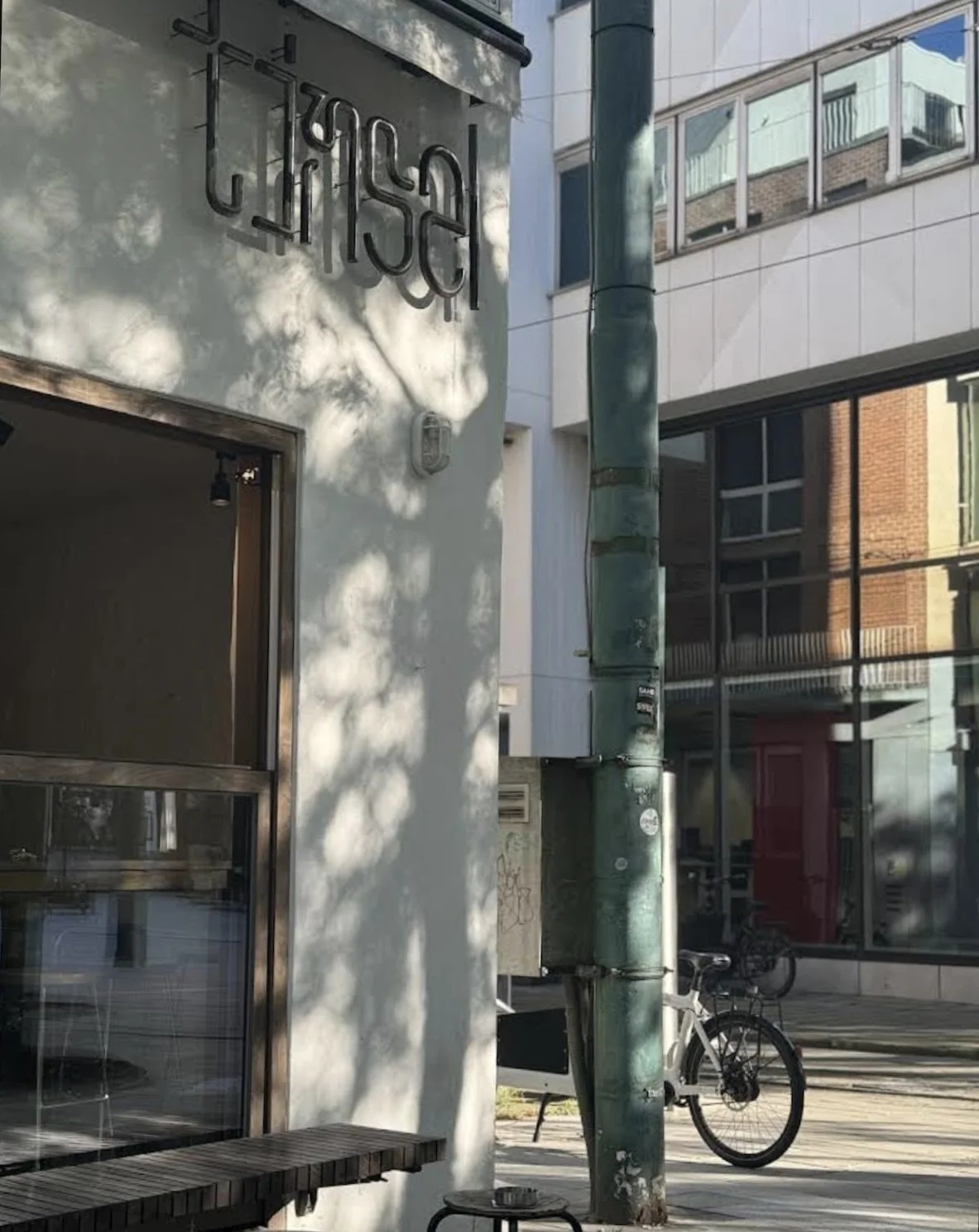A building with a mural and a large window, a street pole, a bicycle, and some buildings reflected in glass, with trees casting shadows.
