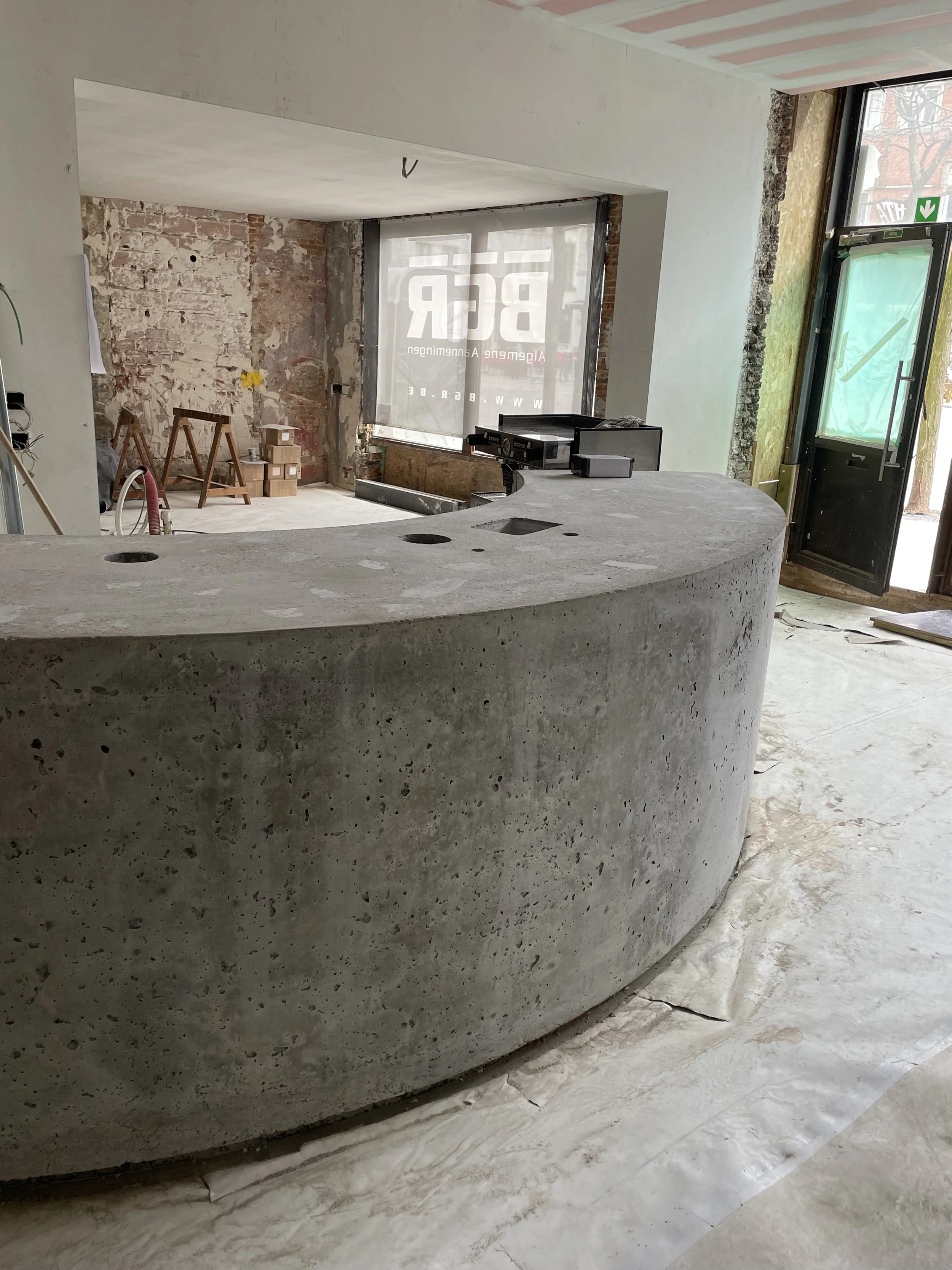 Interior of a building under construction featuring a curved concrete reception or countertop with visible veins and holes, an unfinished wall, and large windows with transparent film covering.