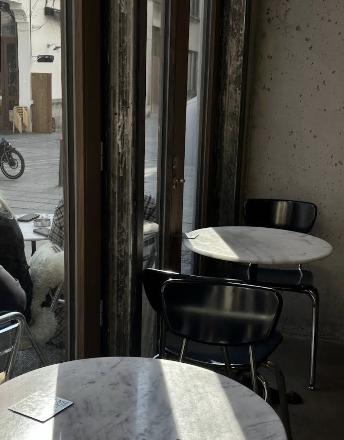 Interior of a cafe with black chairs, white marble tables, a glass door, and a dog sitting outside.