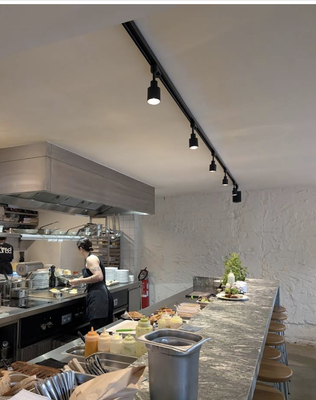 Contemporary open kitchen in a restaurant with chefs preparing food, a marble counter with ingredients, and overhead black track lighting.