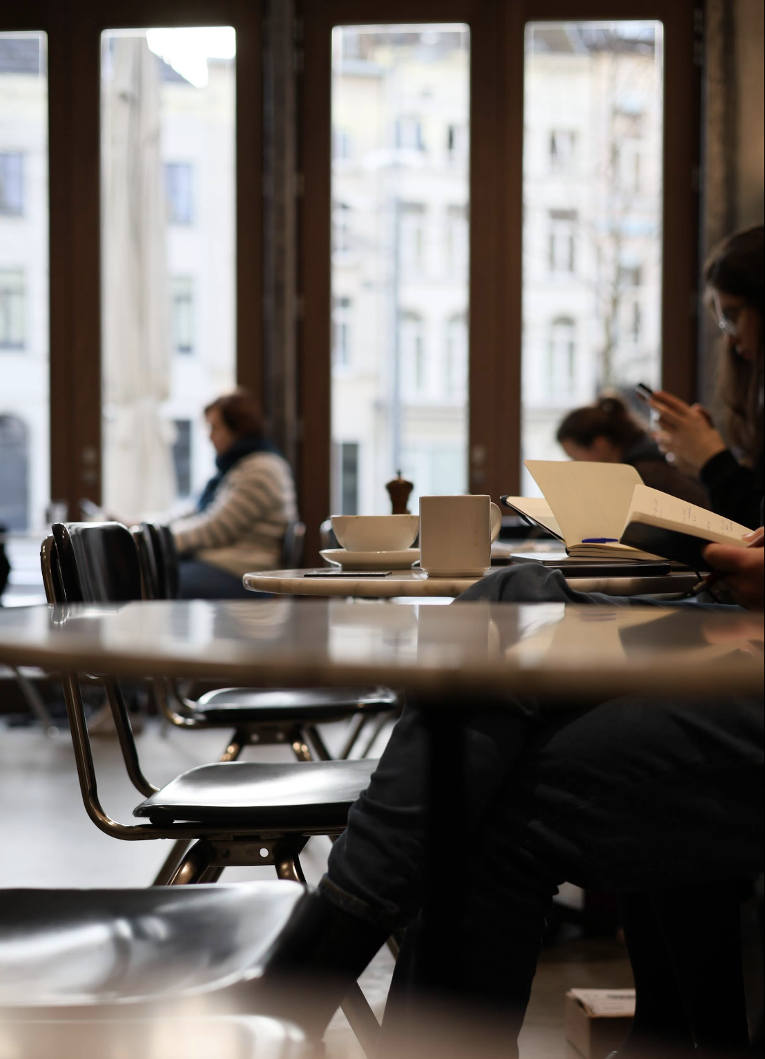Interior of a coffee shop with tables, chairs, and people sitting, some reading, and a large window with a view of buildings outside.