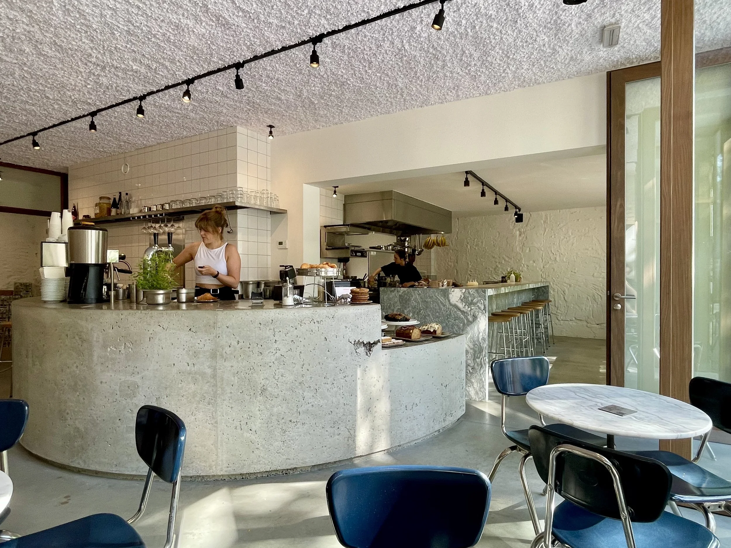 Interior of a modern coffee shop or cafe with a curved stone counter, barstools, and seating area, two women working behind the counter, exposed white brick wall, and large windows letting in natural light.