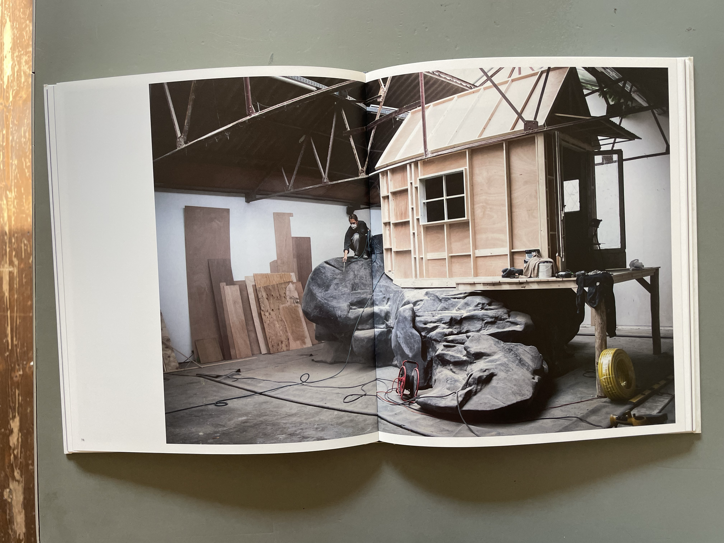 Open pages of a book displaying a photograph of a construction site. The photo shows a partially built wooden structure with a sloped roof, a person wearing a face mask working nearby, and various construction materials and tools scattered around.