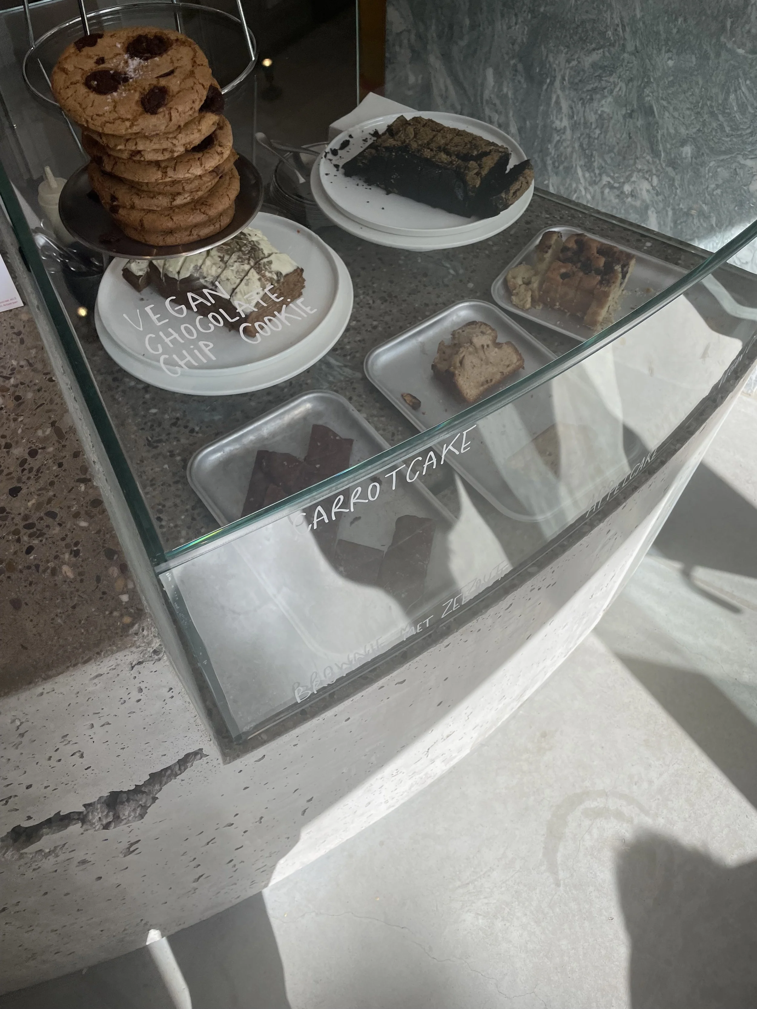 Display case with vegan chocolate chip cookies on a white plate, a slice of carrot cake, a chocolate roll cake, and a tray with a partially eaten baked good, all behind glass with handwritten labels.