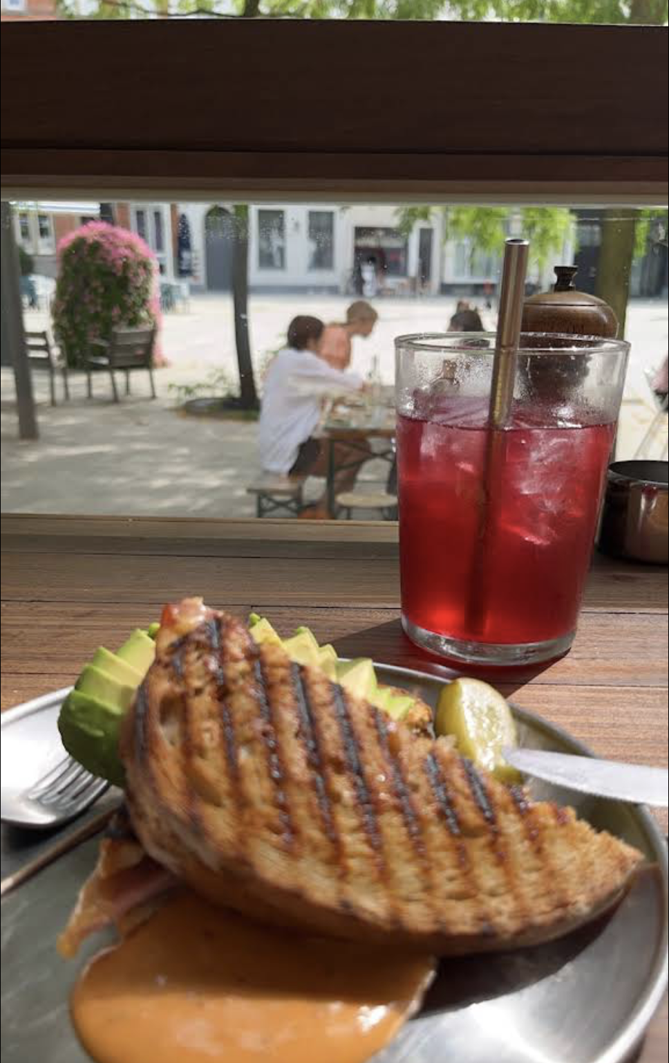 Grilled fish fillet with avocado slices and lemon wedge on a plate, a tall glass of red iced drink with a straw, taken from inside a restaurant or café, overlooking an outdoor patio with people sitting at tables and pink flowers in the background.