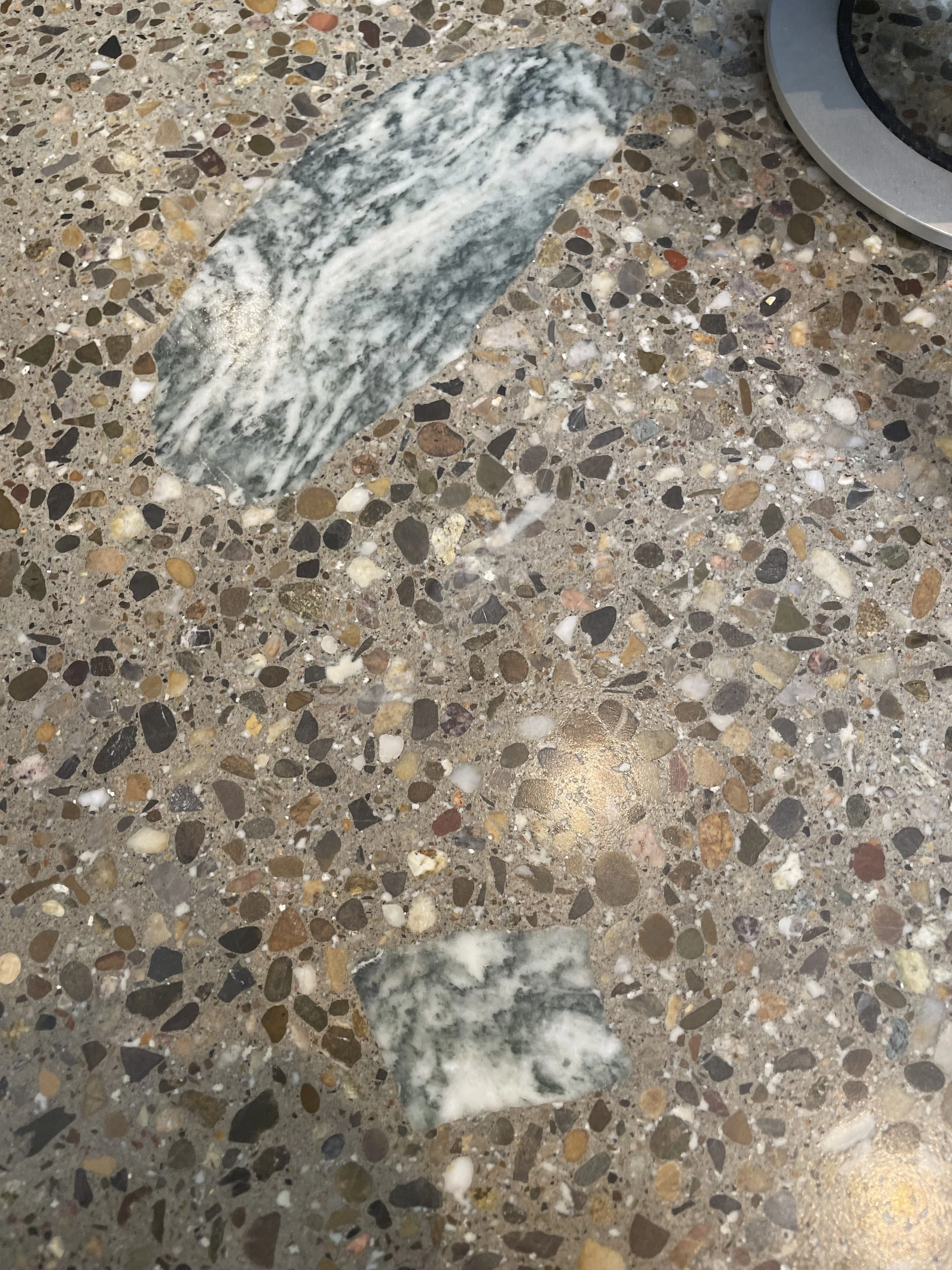 A piece of polished green and white marble on a speckled terrazzo surface, partly beneath a metallic object.