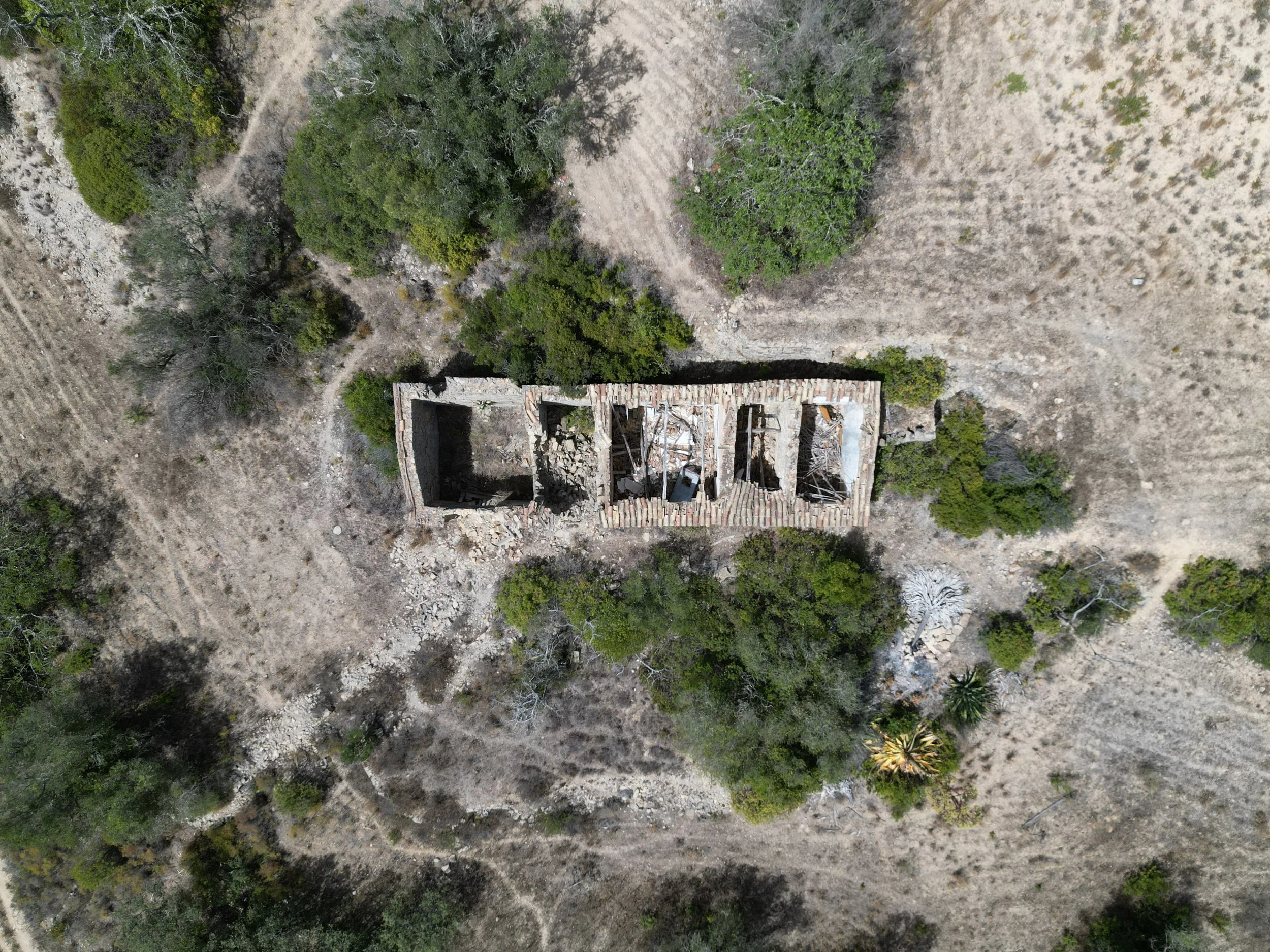 Aerial view of a barren, dry landscape with sparse shrubs and a few green trees, featuring a partially ruined stone building with a tiled roof.
