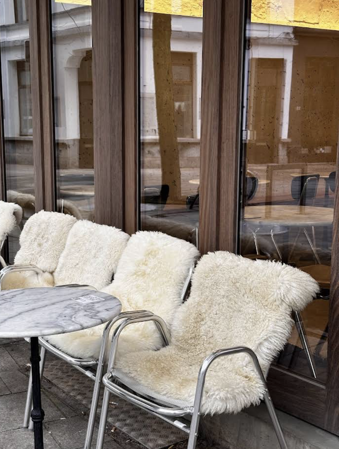 A patio with white fluffy sheepskin-covered chairs and a small round marble table outside a building with wooden and glass walls.