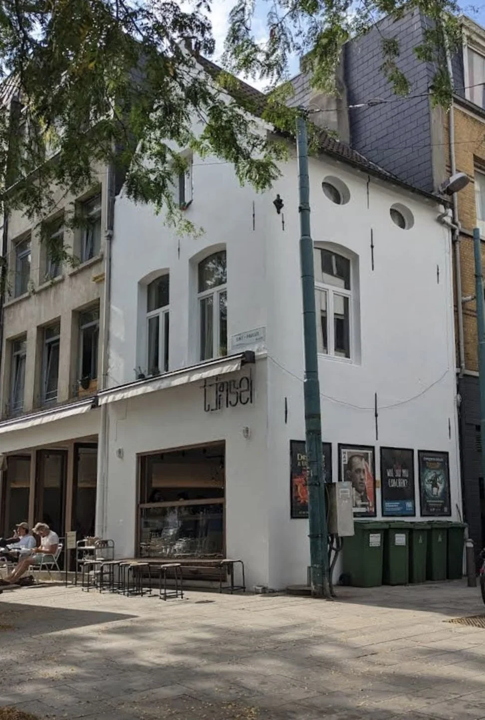 Street view of a white building with large windows, posters outside, green trash bins, and people sitting at tables. A tree partially covers the building.