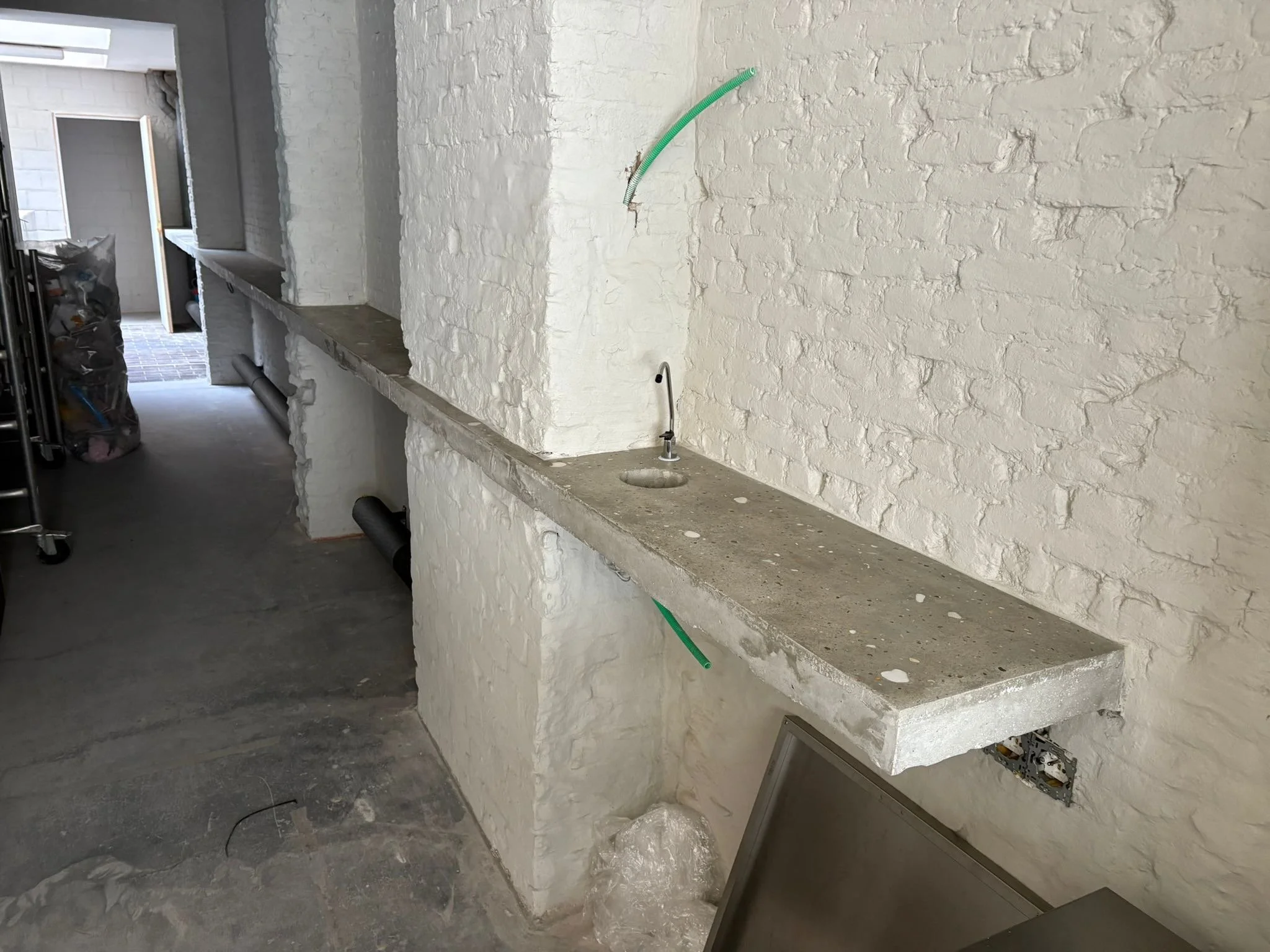 A partially constructed kitchen or utility room featuring white painted brick walls, a concrete countertop with a sink, and construction materials and tools in the background.